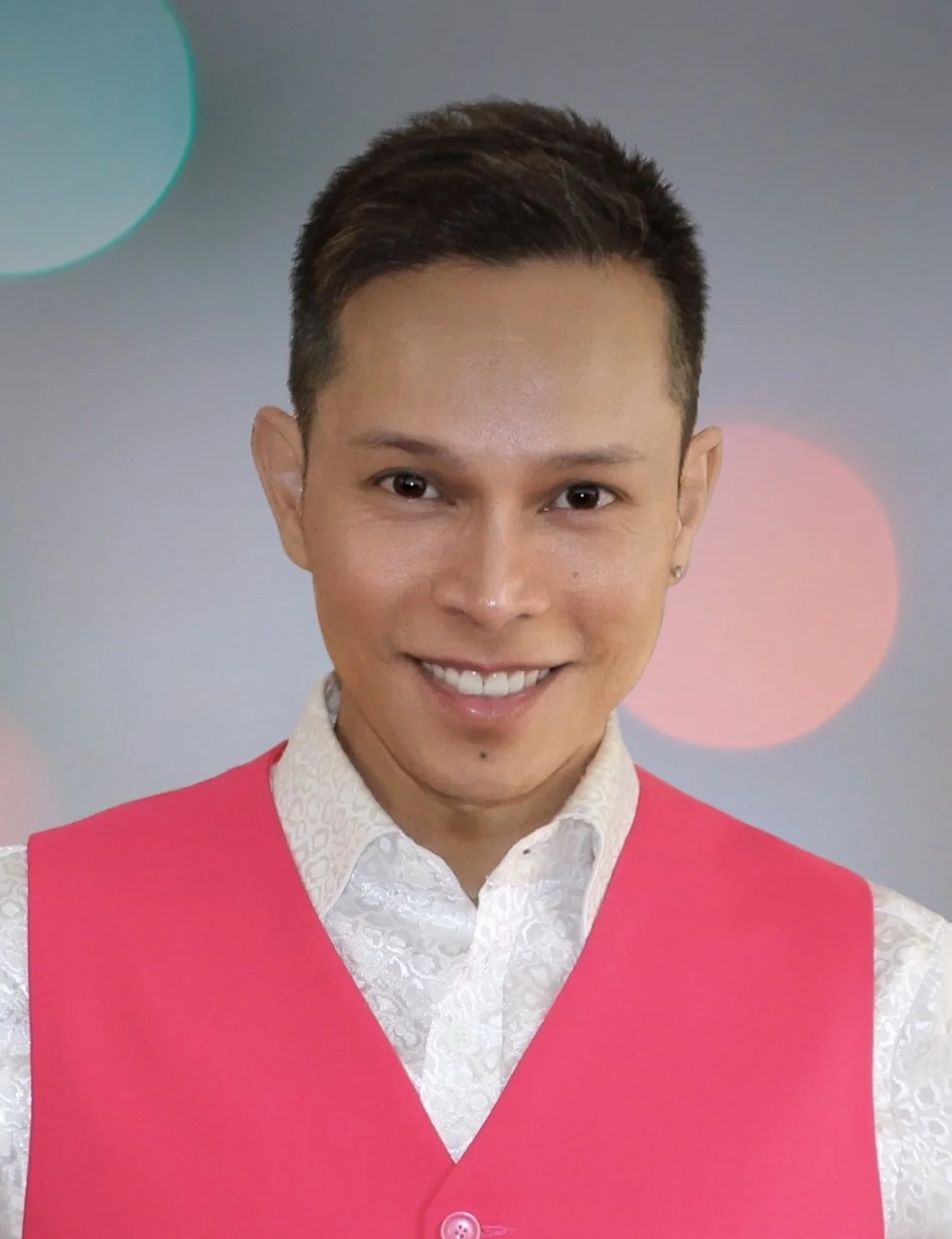 A smiling person with short dark hair wearing a white patterned shirt and a pink vest, standing against a blurred background with colorful bokeh lights.