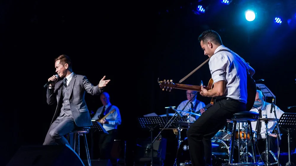 Male singer in a suit performing on stage with a band, including a guitarist and drummer, under blue stage lights.