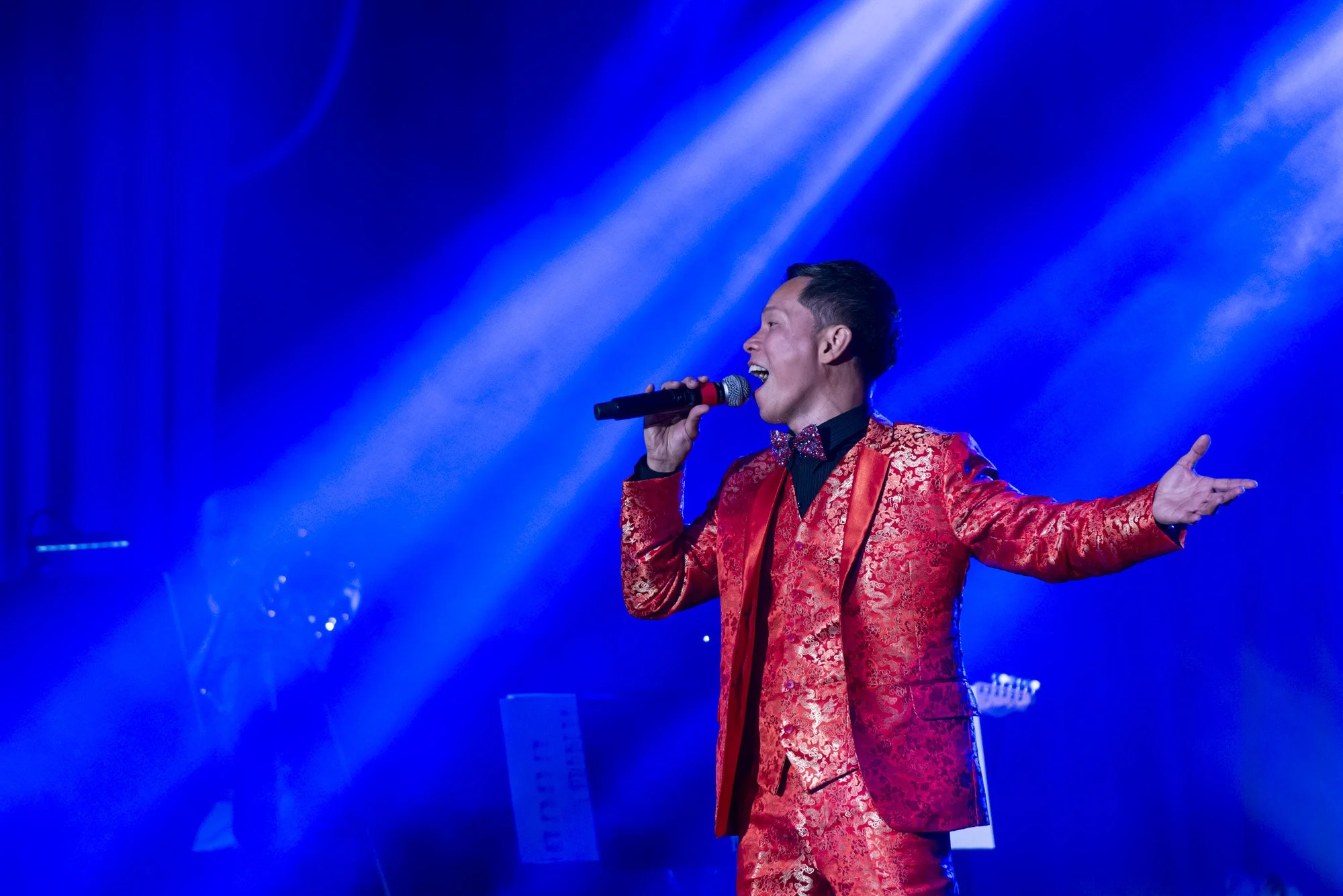 A man in a red patterned suit singing into a microphone on stage under blue lights.