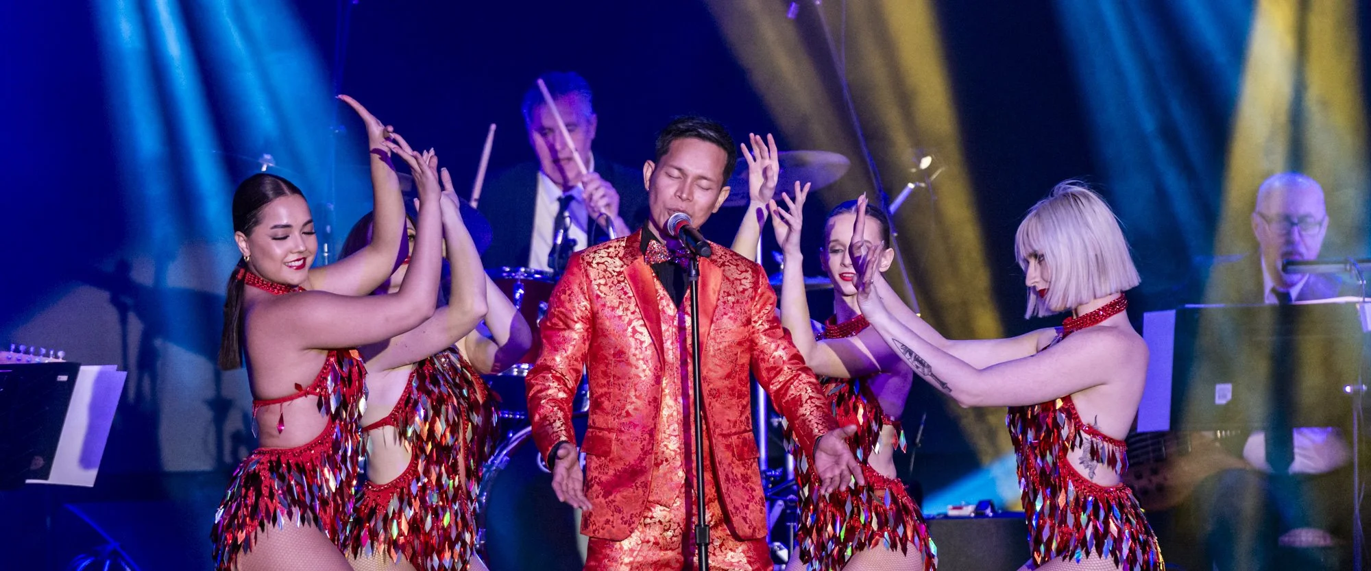 Performer in a red patterned suit singing into a microphone on stage, surrounded by dancers in colorful costumes, with band members playing instruments in the background.