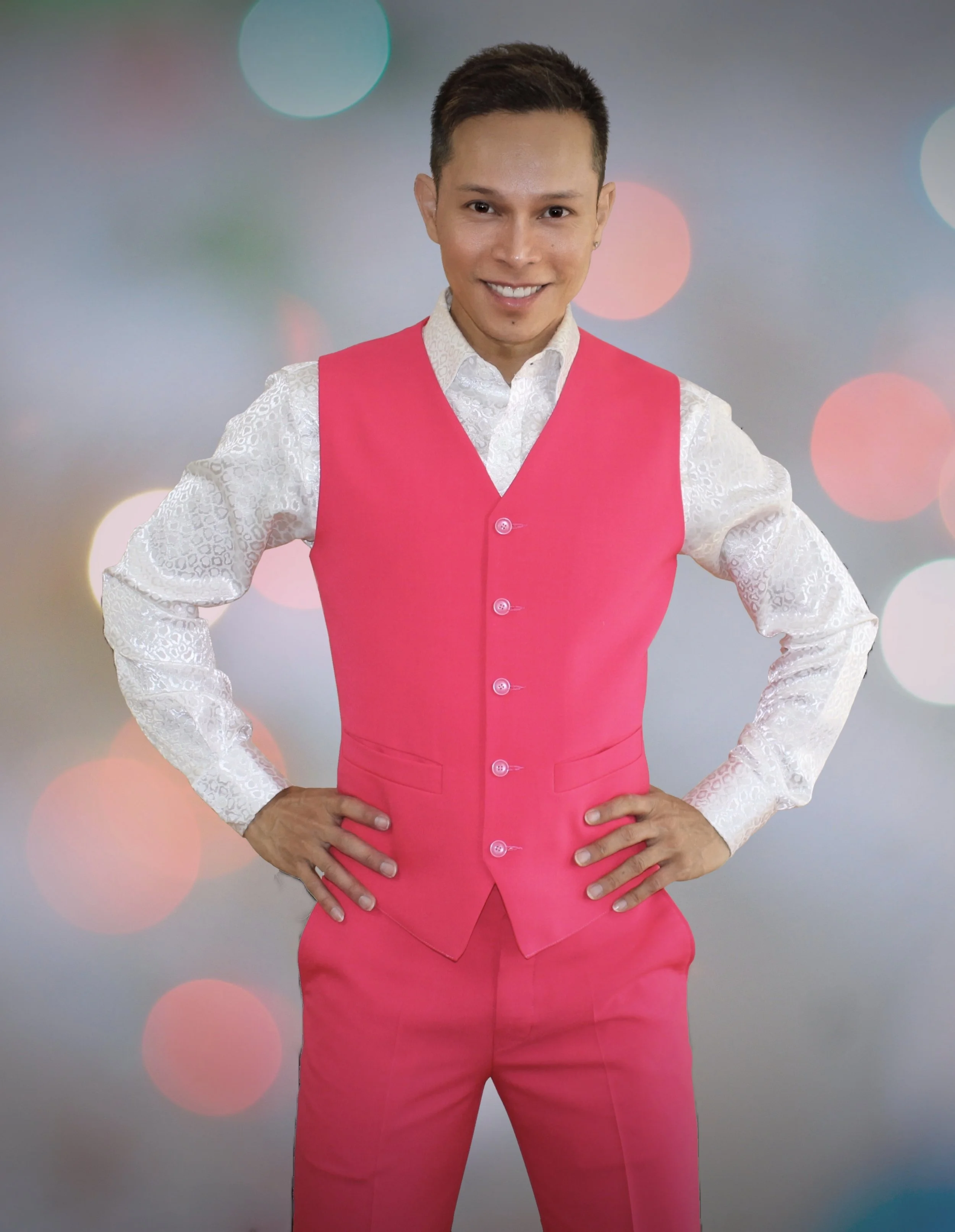 Person wearing a white shirt with a lace pattern and a bright pink vest and pants, standing with hands on hips in front of a colorful blurred bokeh background.