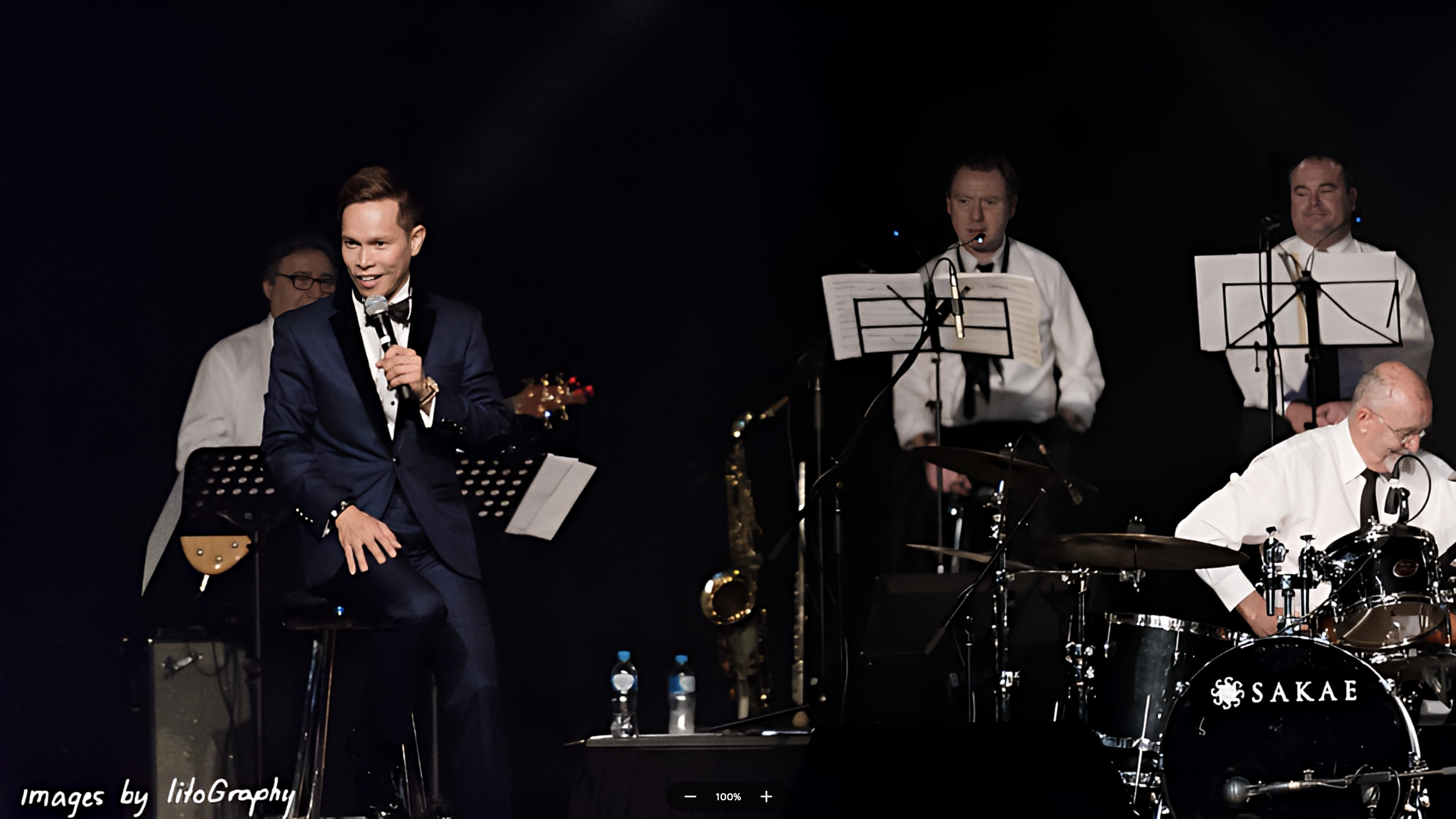 A jazz band performing on stage with four male musicians, including a singer in a navy suit, three men playing instruments, and music stands with sheet music.