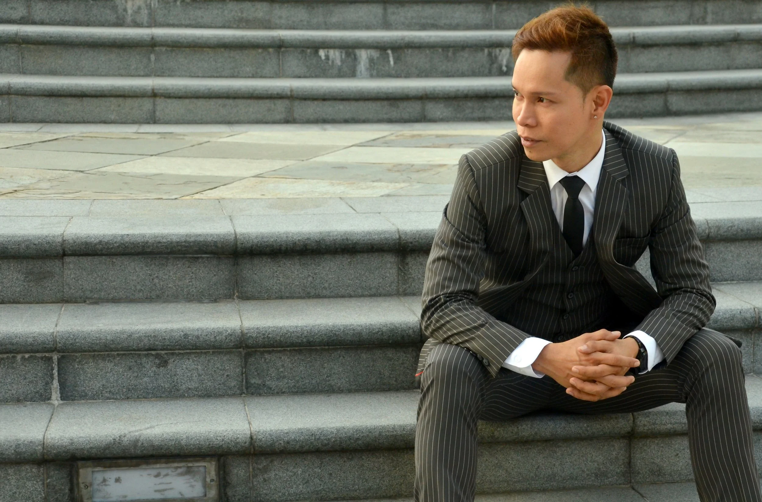 A young man in a black pinstripe suit sitting on stone steps outdoors, looking to his right with hands clasped.