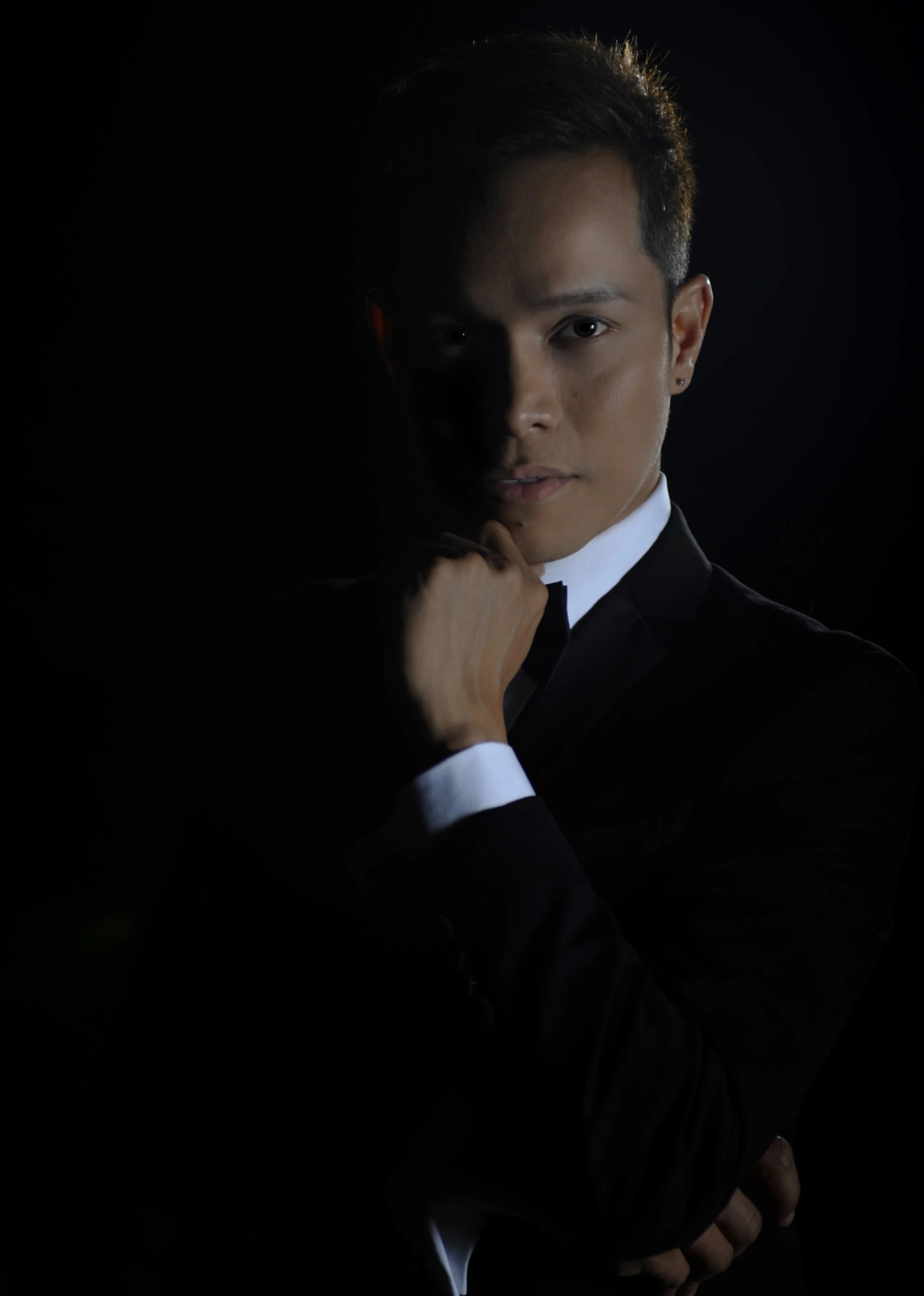 Portrait of a young man in a tuxedo with one hand resting on his chin, illuminated against a black background.