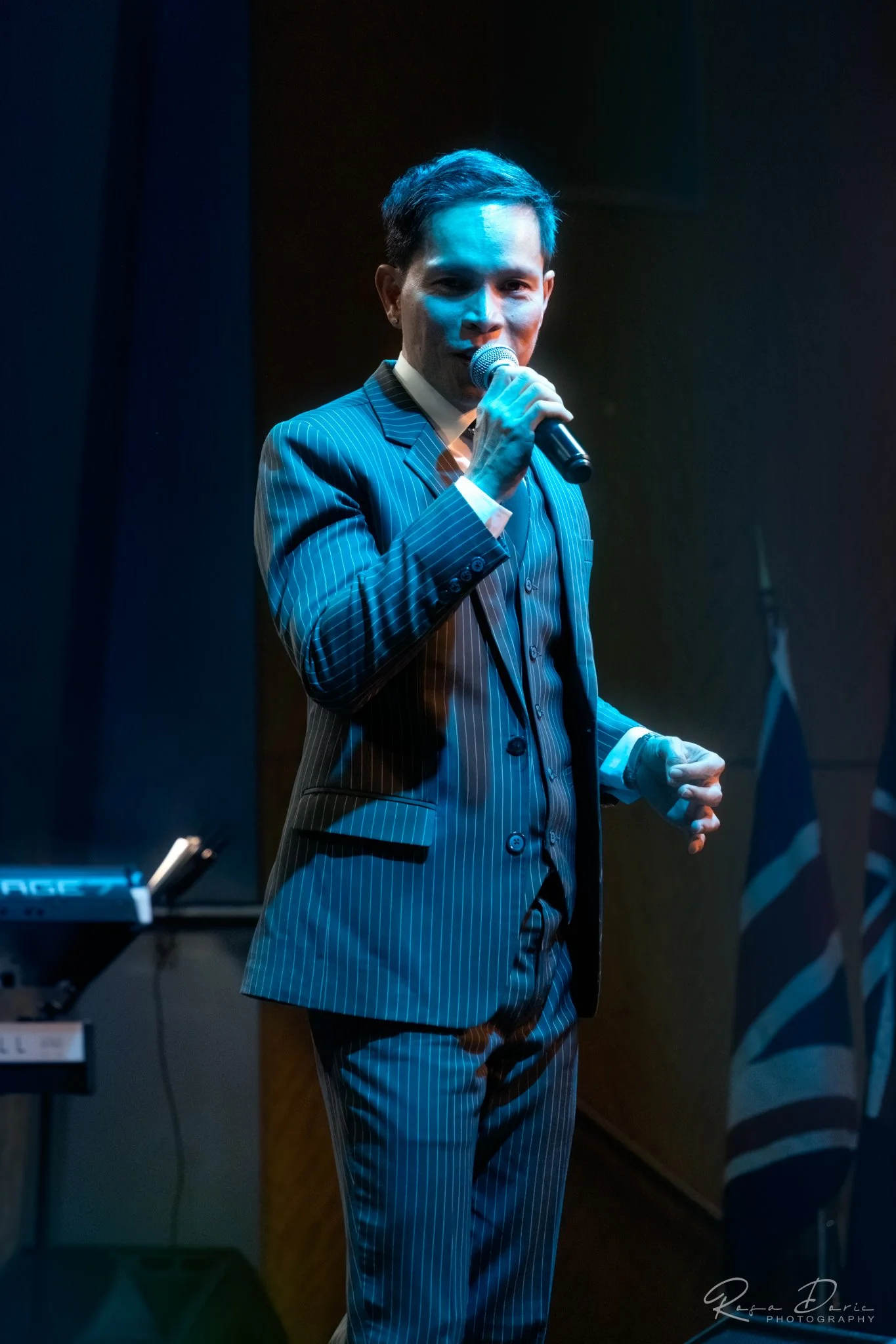 A man in a pinstripe suit speaking into a microphone during a performance or event, with a keyboard visible to the left and a dark background.