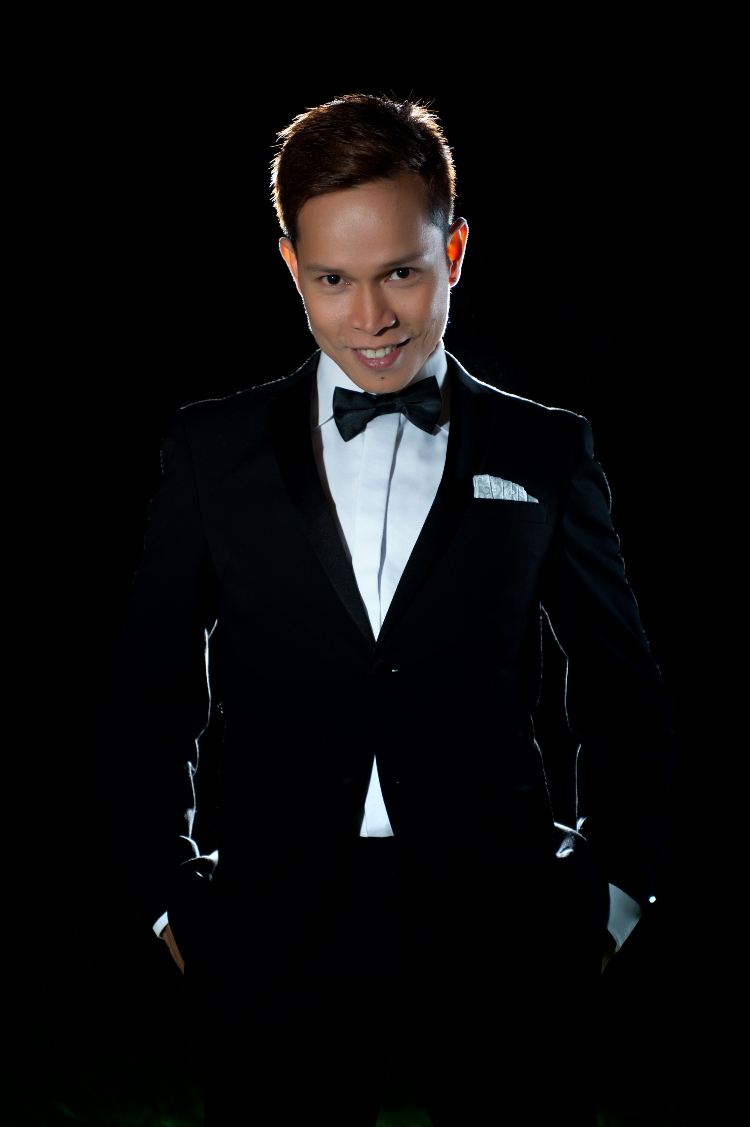 Portrait of a young man in a tuxedo with a bow tie, smiling, with a dark background.