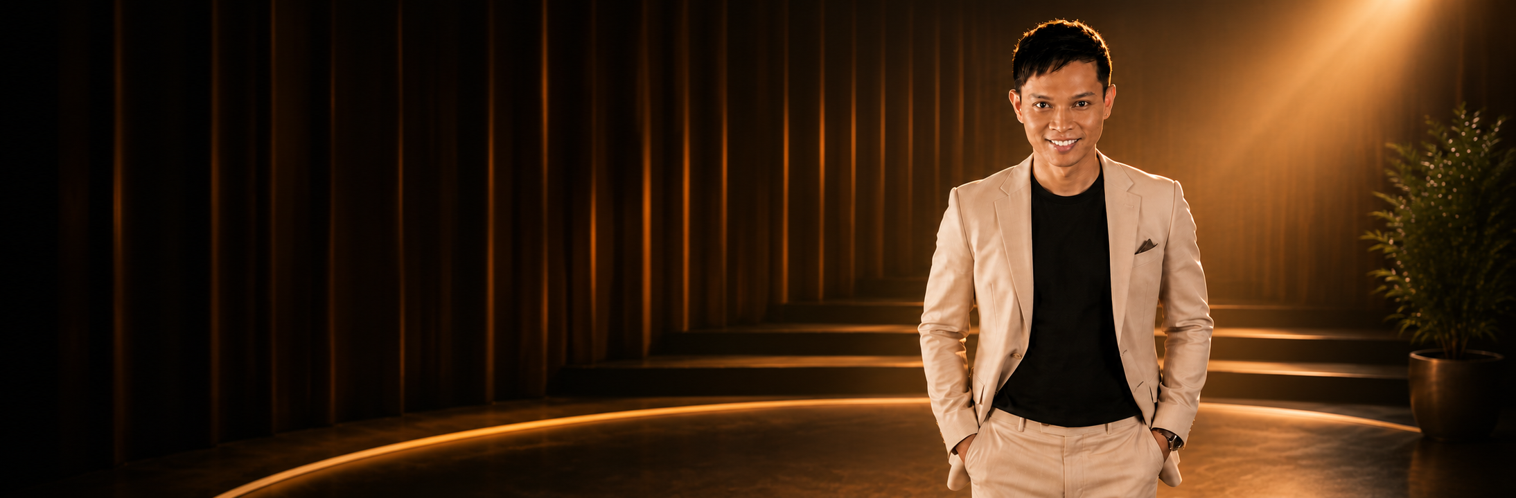 A man dressed in a tan suit and black shirt stands in a dimly lit, modern interior with wooden wall panels and warm lighting.