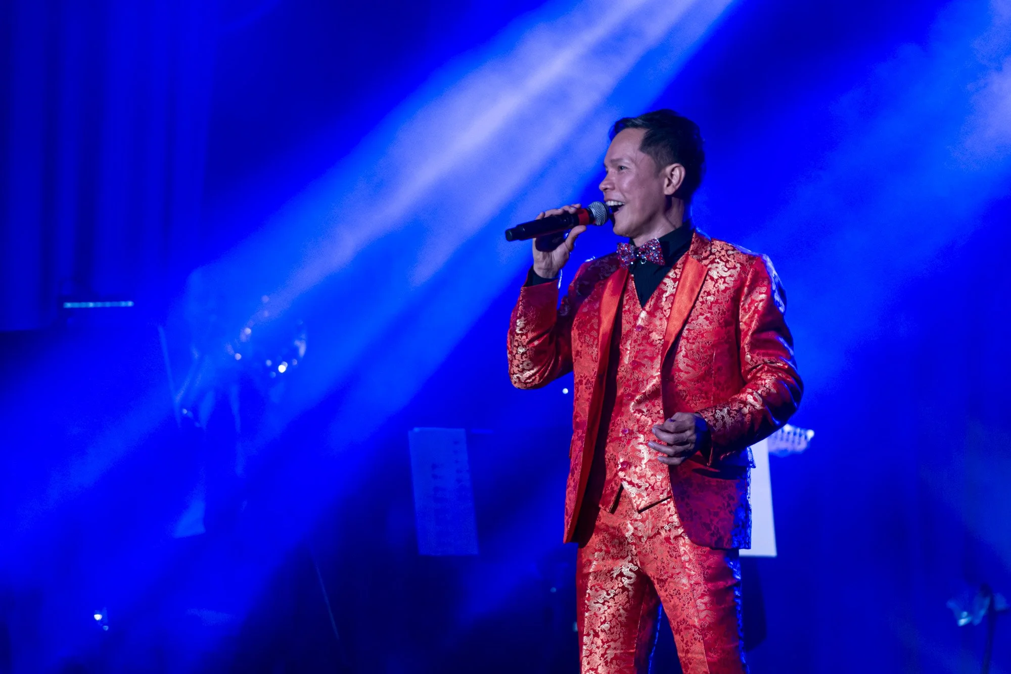 A man in a red, patterned tuxedo with a bow tie is singing into a microphone on stage, illuminated by blue stage lighting.