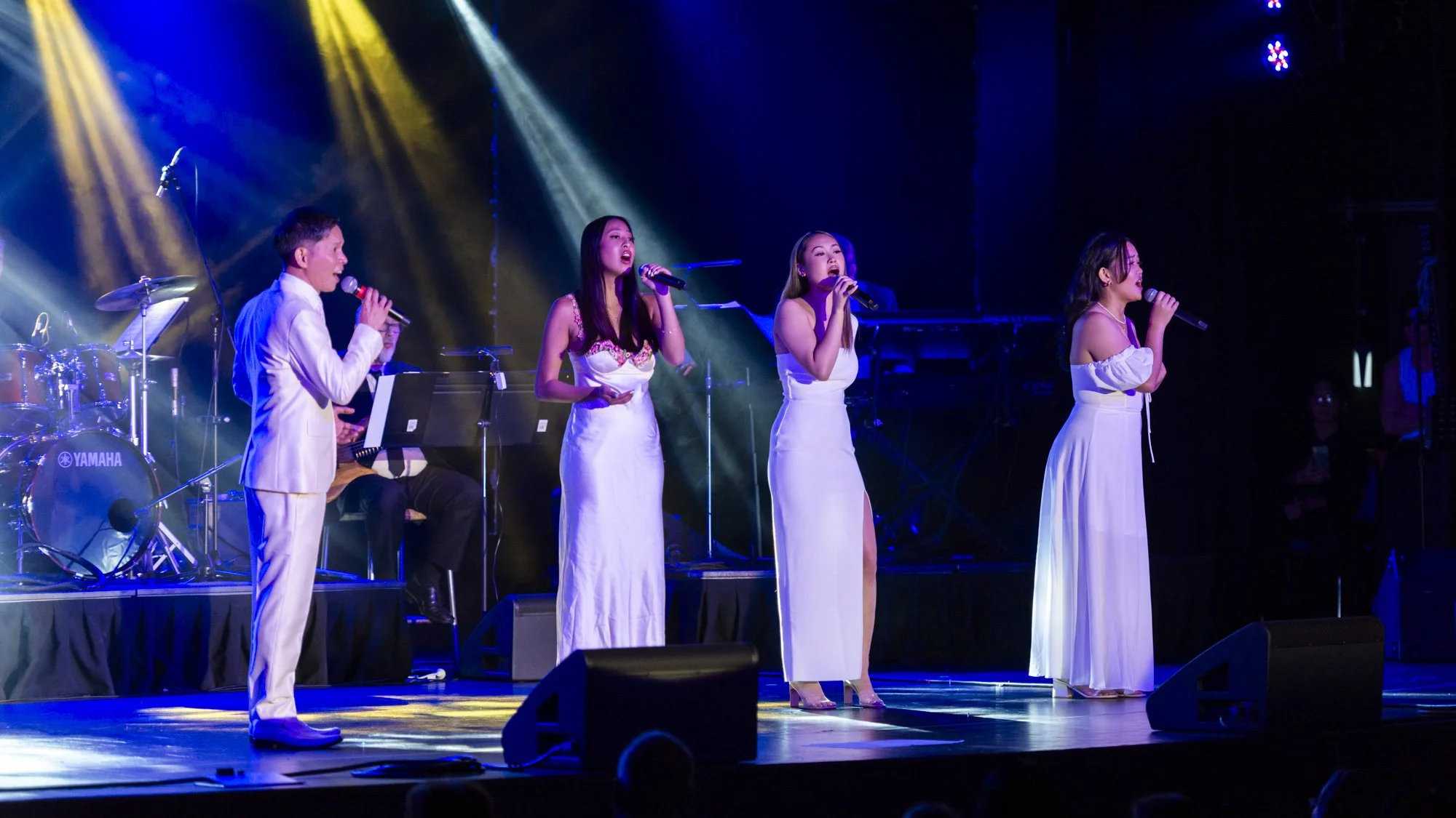 A live musical performance on stage with four female singers in white dresses and one male singer in a white suit, accompanied by a band with drums and keyboard, under colorful stage lighting.