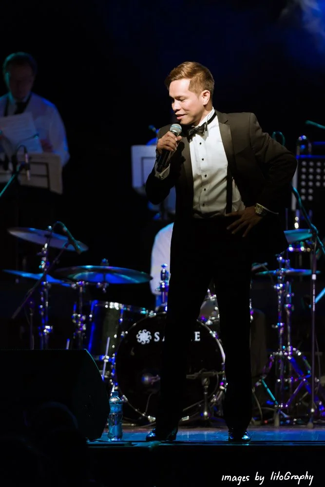 Man in a tuxedo singing into a microphone on stage with a band in the background.