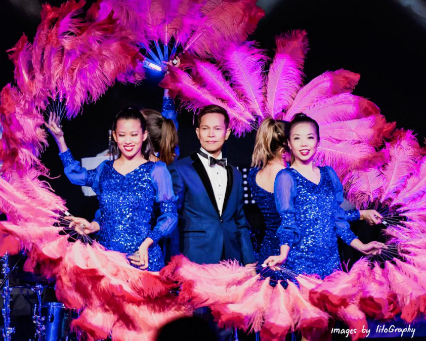 Performer in blue sequined dress holding pink feathers, standing beside man in tuxedo with bow tie, on stage with a backdrop of pink feathers and stage lights.