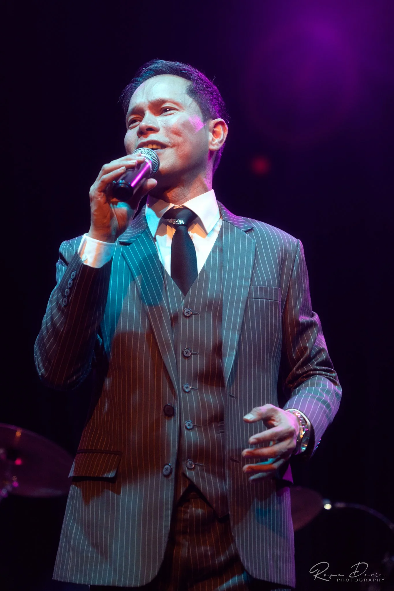 A man in a pinstripe suit holding a microphone and singing on stage under purple lighting.