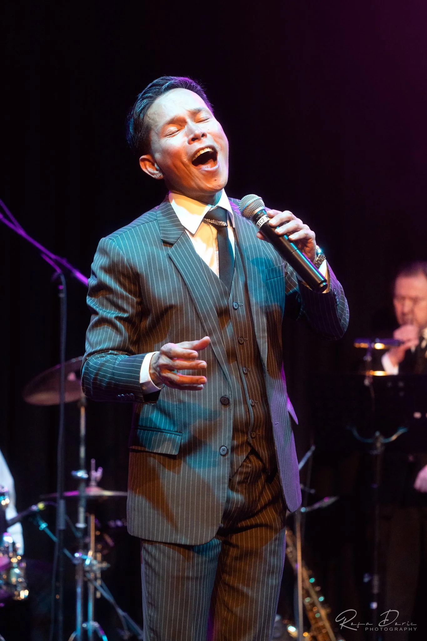 A man in a pinstripe suit sings passionately into a microphone on stage with music equipment and a musician in the background.