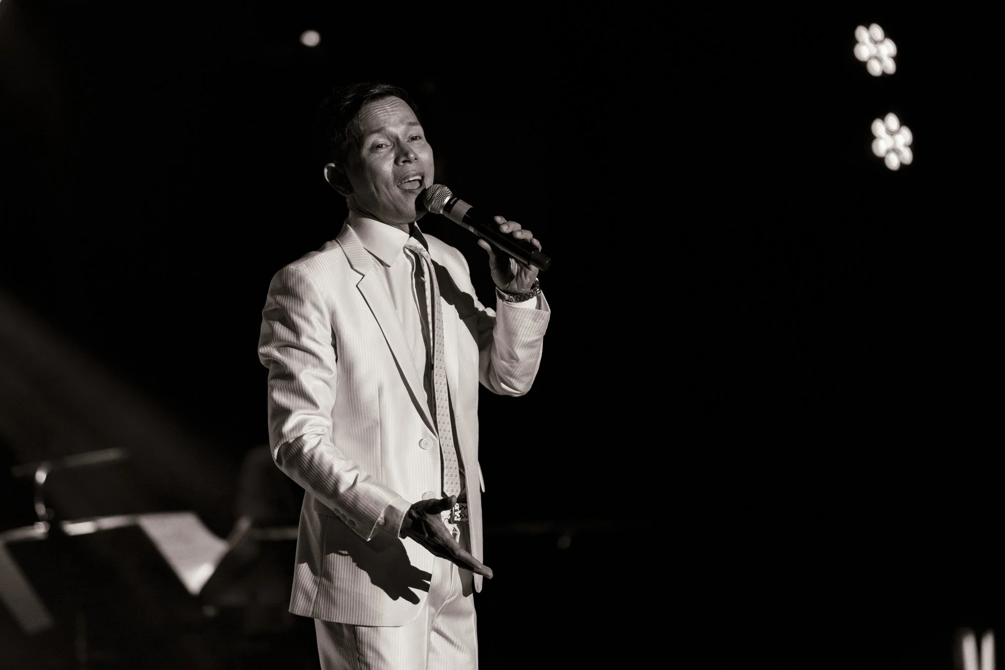 A man in a light-colored suit, holding a microphone, performing on stage with a dark background and bright stage lights.