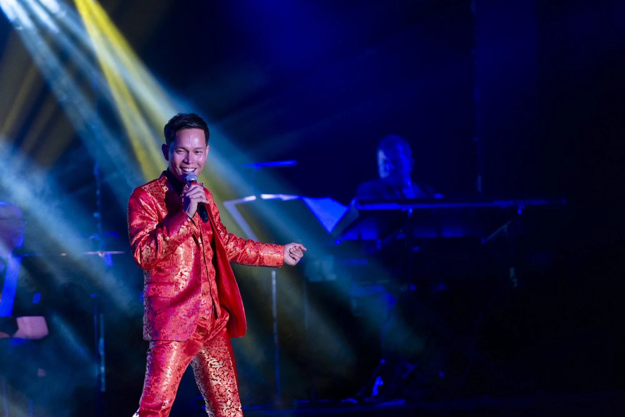 A male performer in a bright red, patterned suit singing into a microphone on stage with colorful lighting and a keyboardist in the background.
