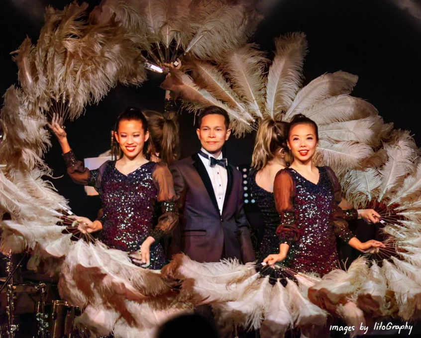 Three performers, two women in sequined dresses and one man in a tuxedo, posing on stage behind large feather fans.