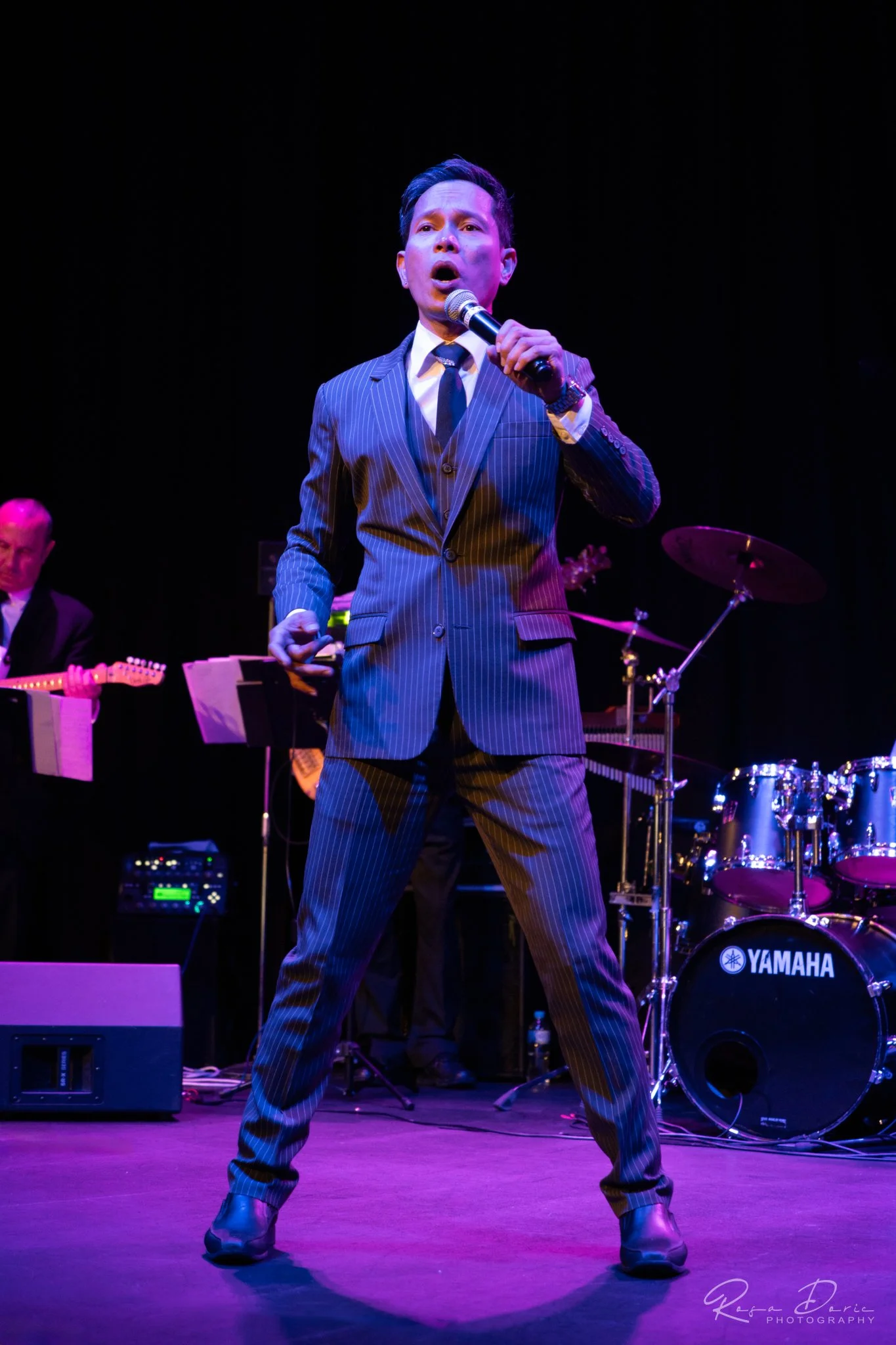 A man in a pinstripe suit singing into a microphone on stage, with musical instruments and a drummer in the background, under colorful stage lighting.