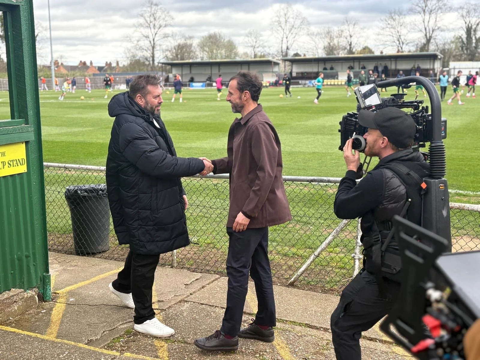 Two men shake hands near a fence at a sports field. One wears a black puffer jacket, the other a brown blazer. A cameraman films them, wearing a black cap, jacket, and carrying filming equipment. In the background, people are playing soccer on the fi