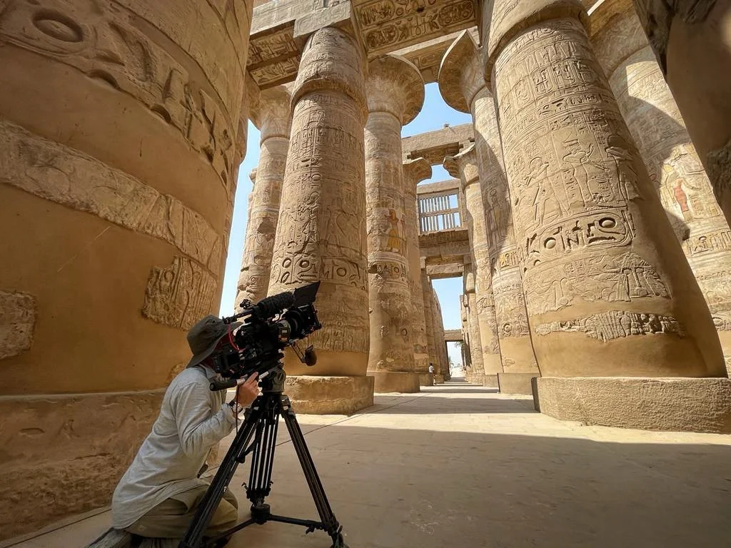 Lorian DOP adjusting a camera on a tripod in front of ancient Egyptian temple columns with hieroglyphics and carvings, under a clear sky.