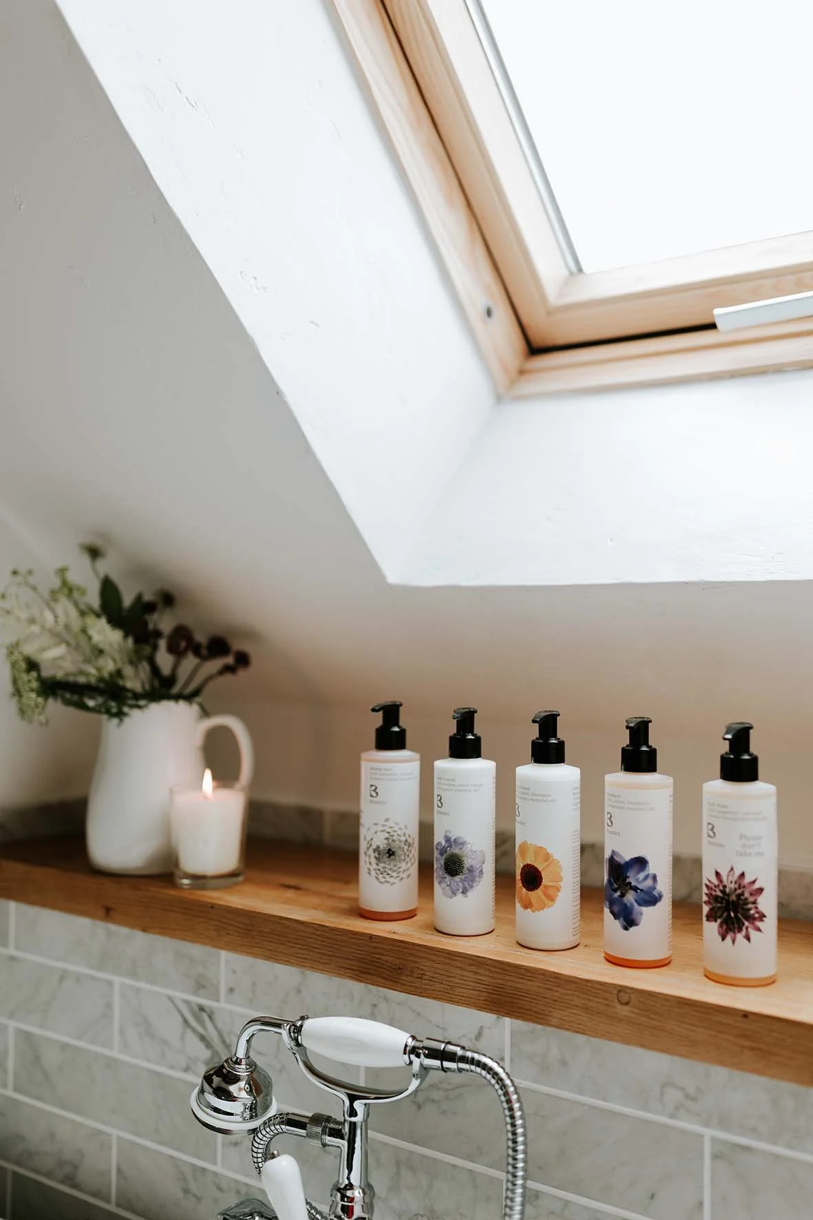 Bathroom shelf with five bottles of skincare products, a white pitcher with flowers, a lit candle, and a bathroom sink faucet.
