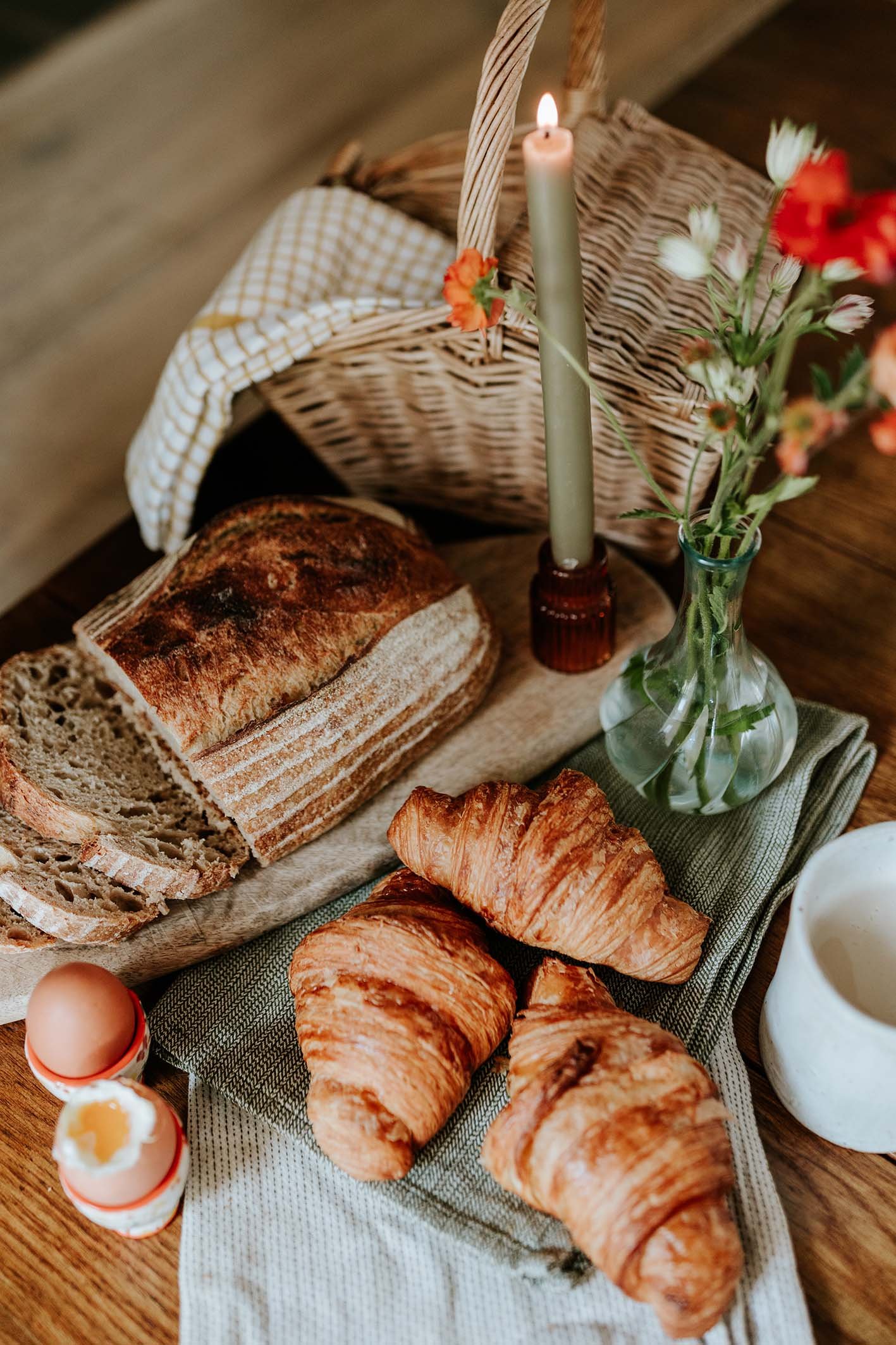 A basket of baked goods including a loaf of bread and croissants, along with eggs in an egg carton, a jar of milk, a lit candle in a brown holder, and a vase with flowers, on a wooden table.