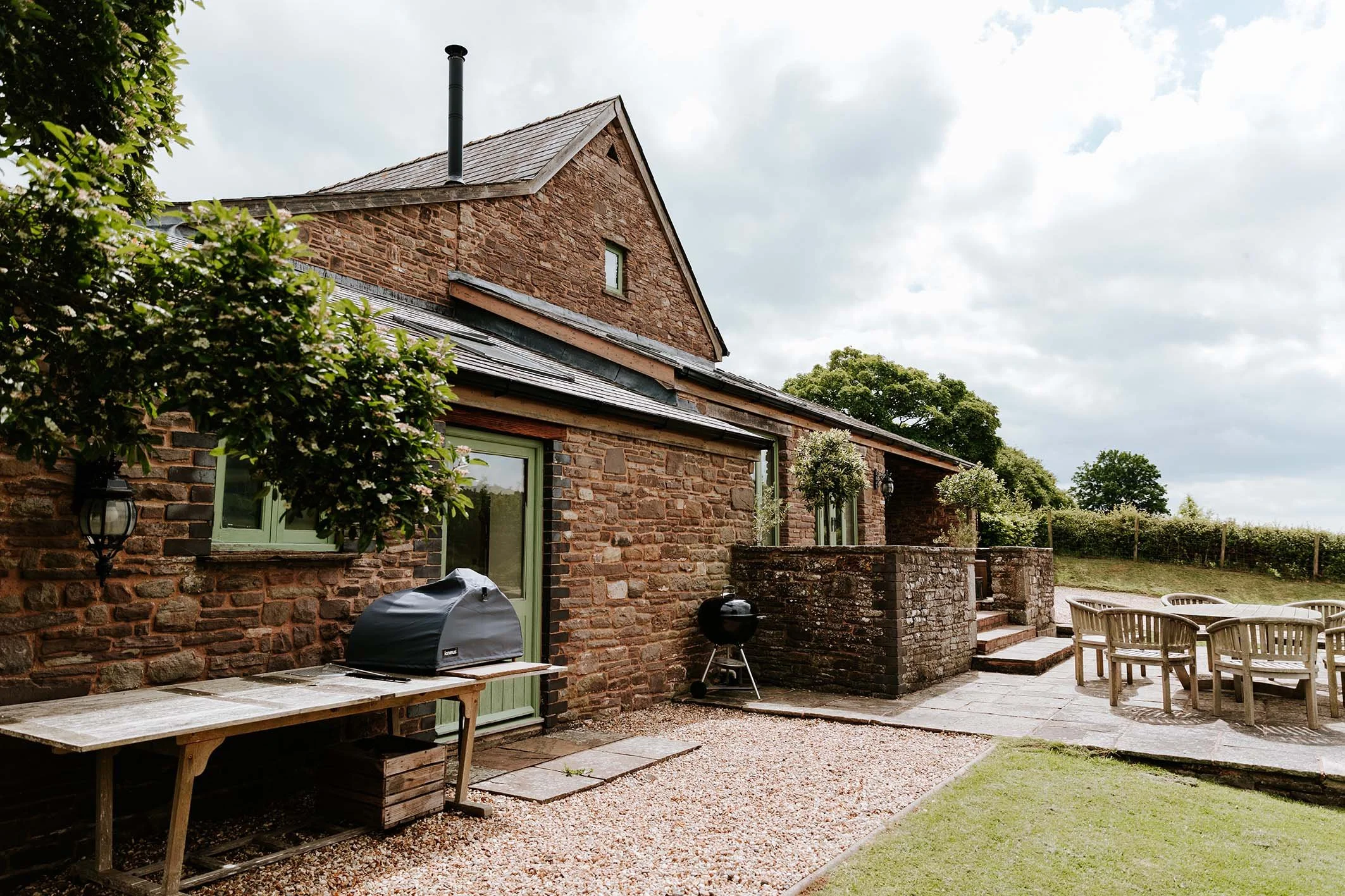 A backyard patio area adjacent to a brick house with a sliding door, featuring outdoor furniture, a barbecue grill, potted trees, and a view of a grassy yard and trees in the background under a cloudy sky.