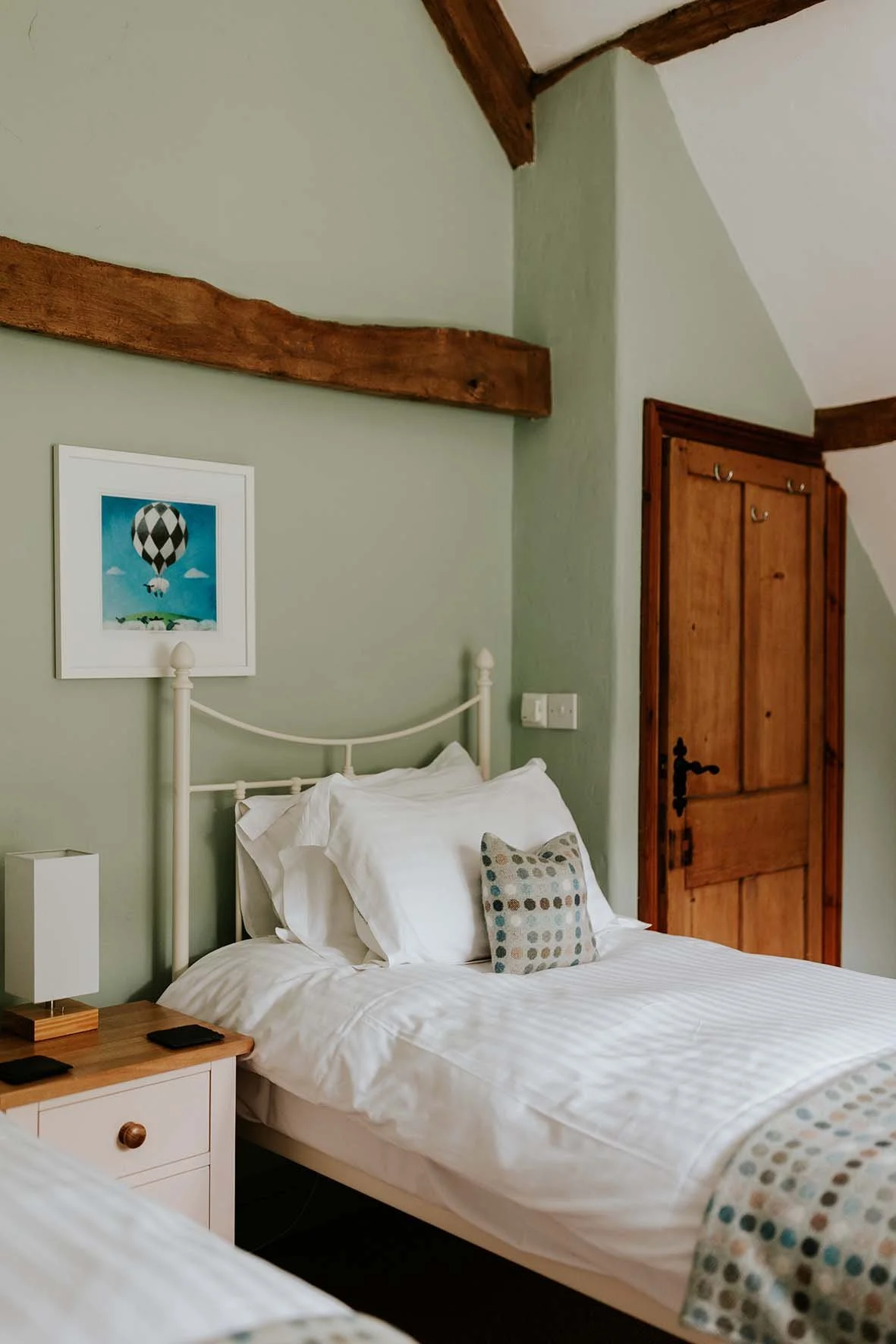 A cozy bedroom with a white bed featuring a decorative pillow, a wooden nightstand with a lamp, a framed picture of a hot air balloon on the green wall, and a wooden door with iron hinges under a sloped ceiling with exposed wooden beams.