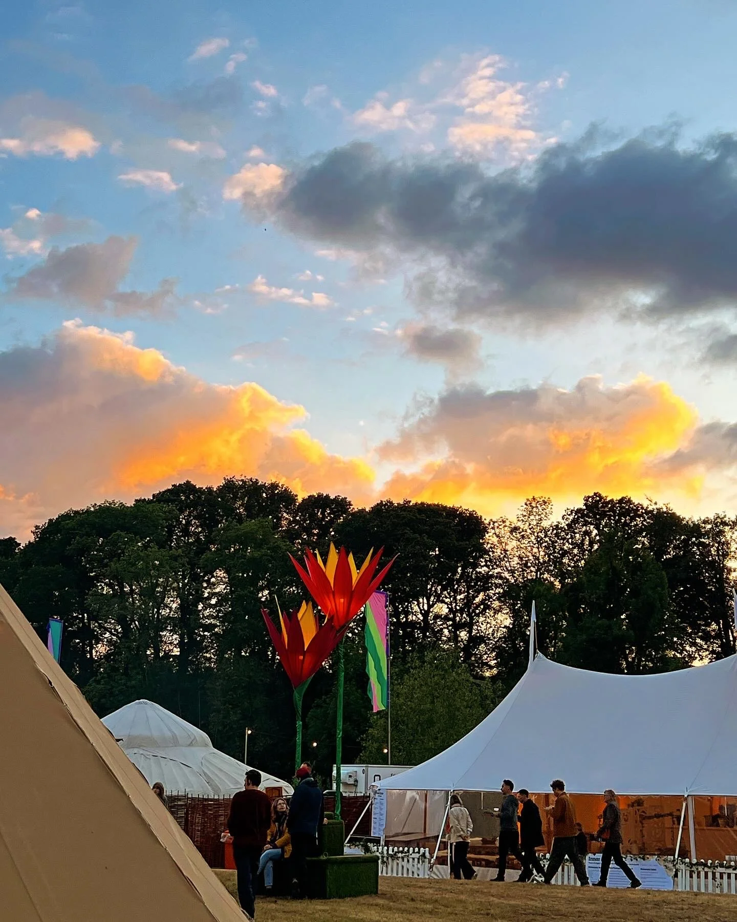 People walking and sitting near large tents and illuminated sculptures resembling oversized tropical flowers, with a backdrop of trees and a colorful sunset sky with clouds.