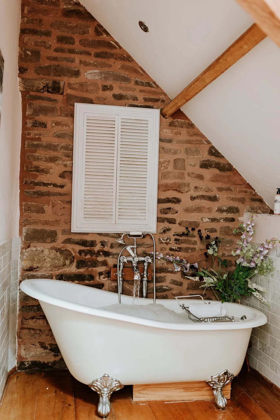 A vintage freestanding bathtub with clawfoot design filled with water, situated against a rustic red brick wall with an arched ceiling. There is a small basket on the tub holding a white towel, and decorative flowers are placed nearby. A white vent o