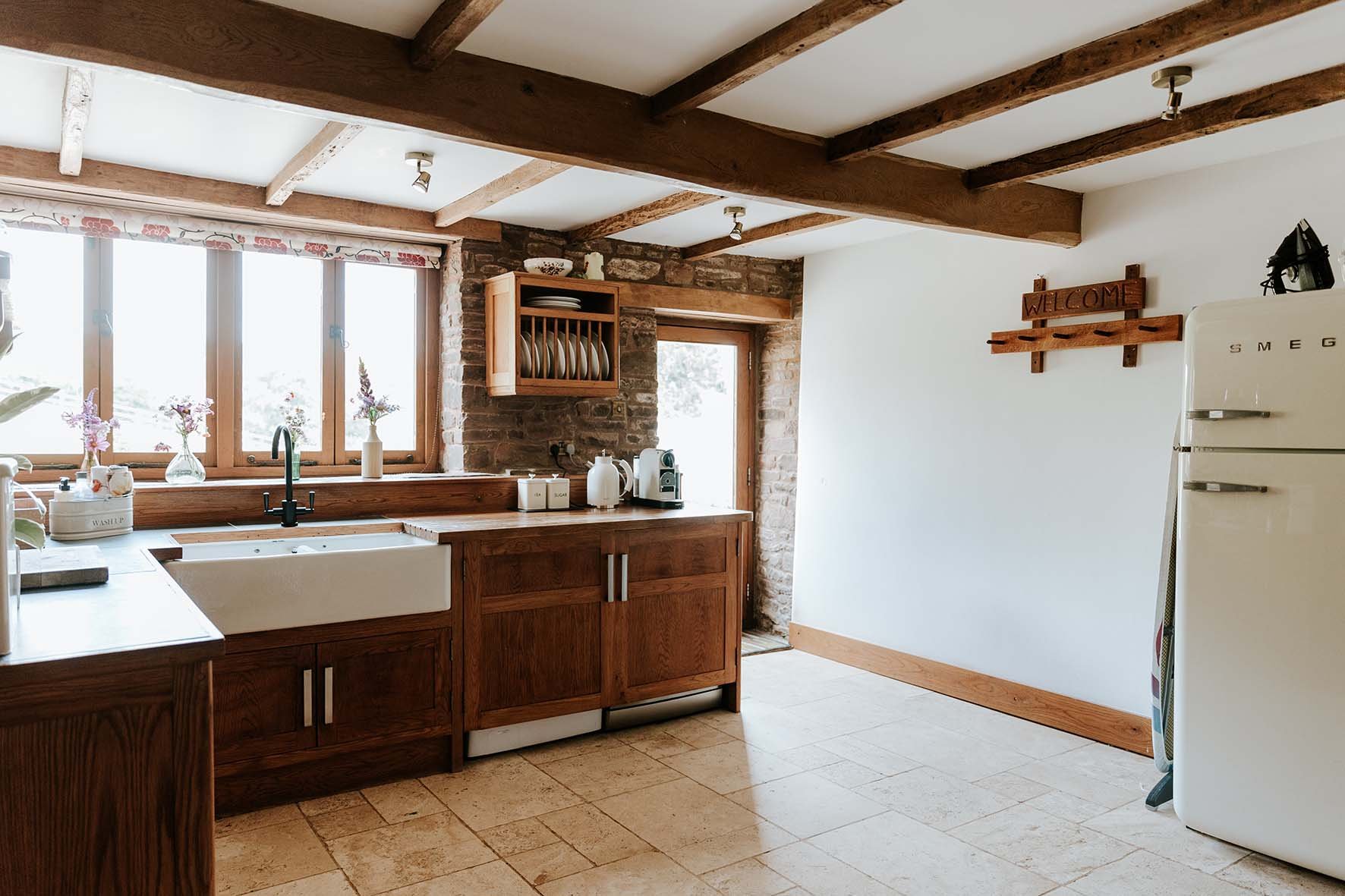 Rustic kitchen with wooden cabinets, a white farmhouse sink under a large window, brick wall accents, and a cream-colored Smeg refrigerator.