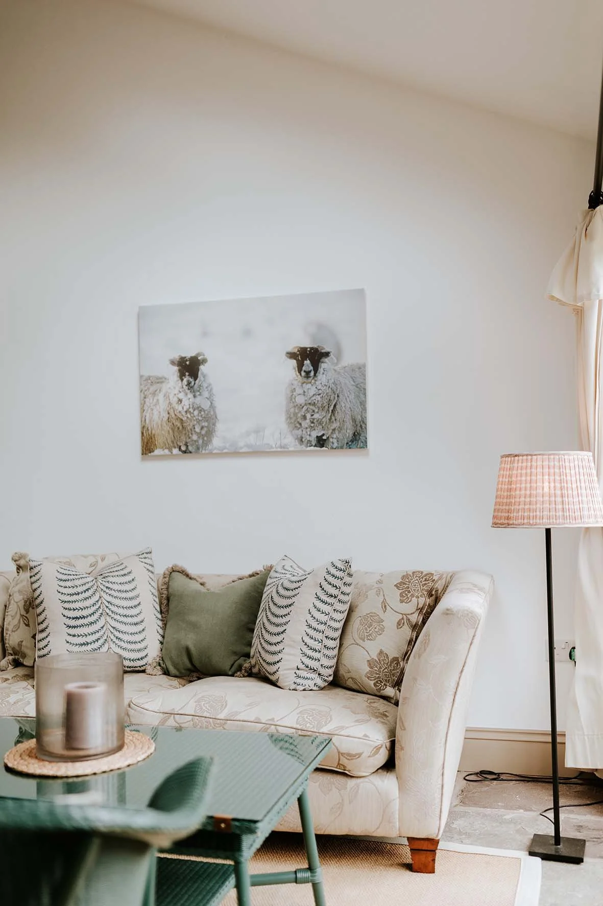 Living room with a floral patterned beige sofa, decorative pillows, a glass coffee table with candles, a pink checkered floor lamp, a window with white curtains, and a framed photo of sheep on the wall.