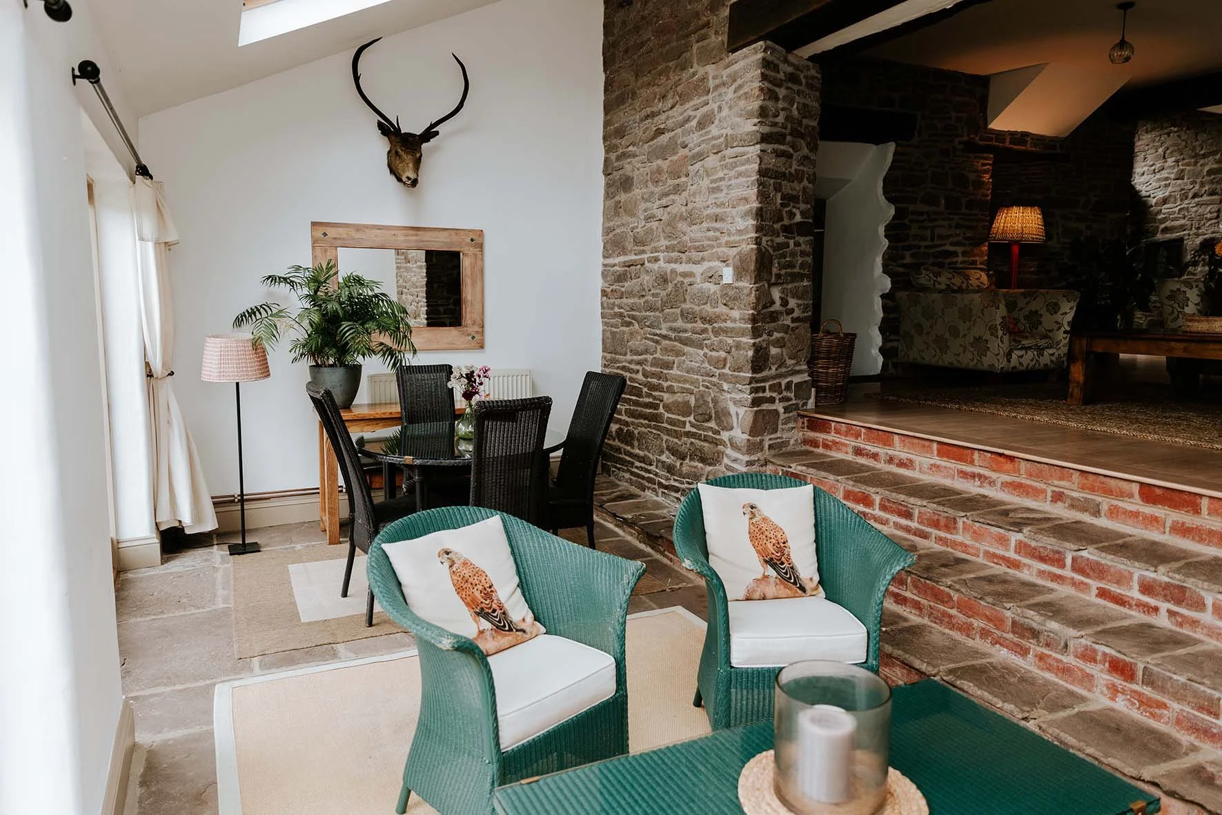 Cozy living and dining area with brick accents, green wicker chairs with bird pillows, a round glass table, a kitchen area with a dining table, and decorative elements like a deer head mounted on white wall, a mirror, plants, and lamps.