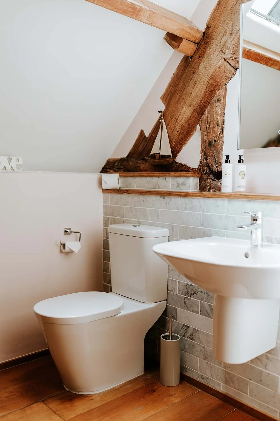 A bathroom with a white toilet, a small white sink, a wall-mounted mirror, and decorative wooden beams. The counter behind the toilet has a model sailboat and two white bottles. There is a wood floor and a partial brick wall.