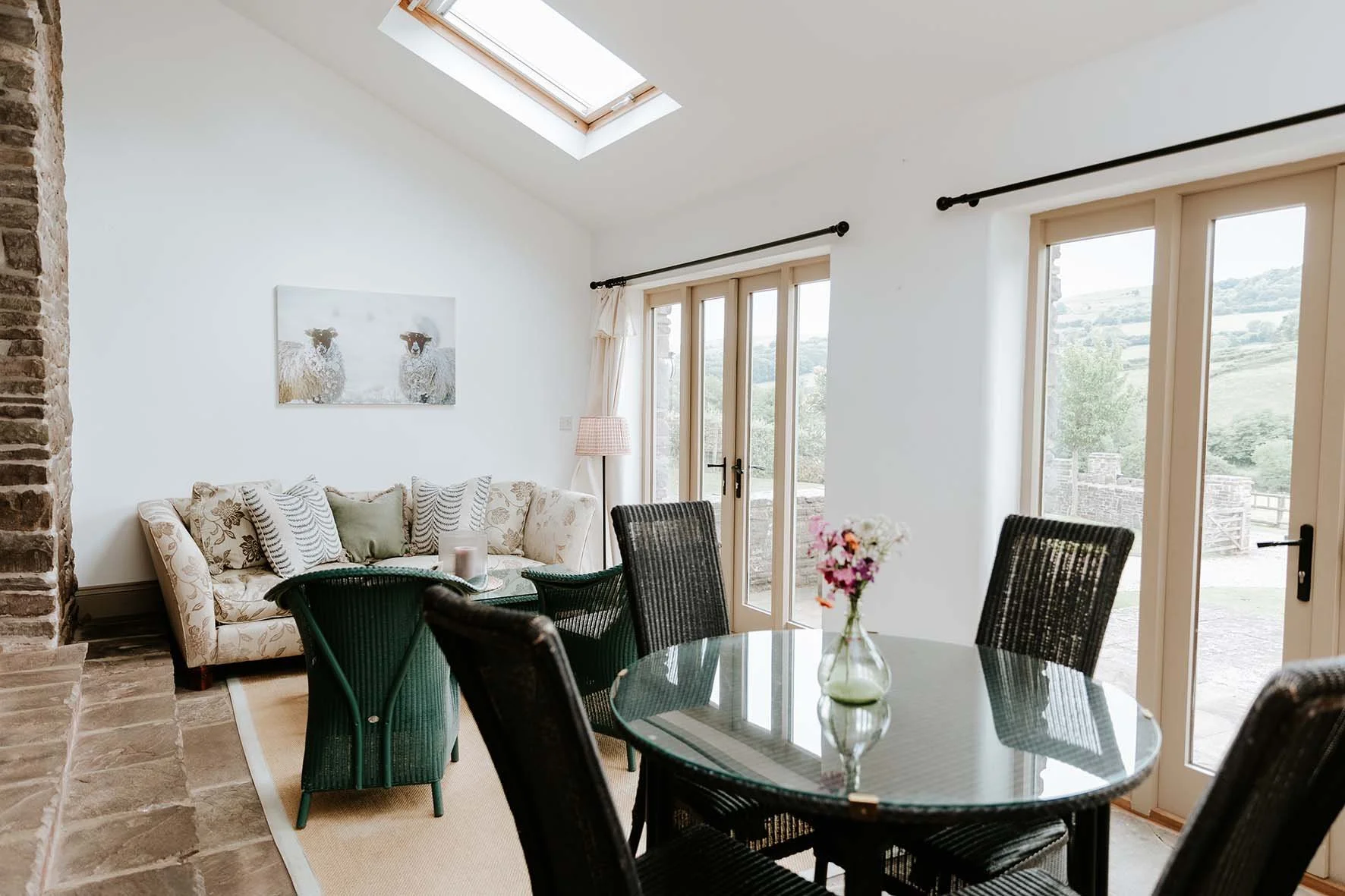 Bright living room with a round glass dining table, black chairs, a cream sofa with patterned cushions, a pink lamp, large windows and a sliding door, a skylight, and a mountain view outside.