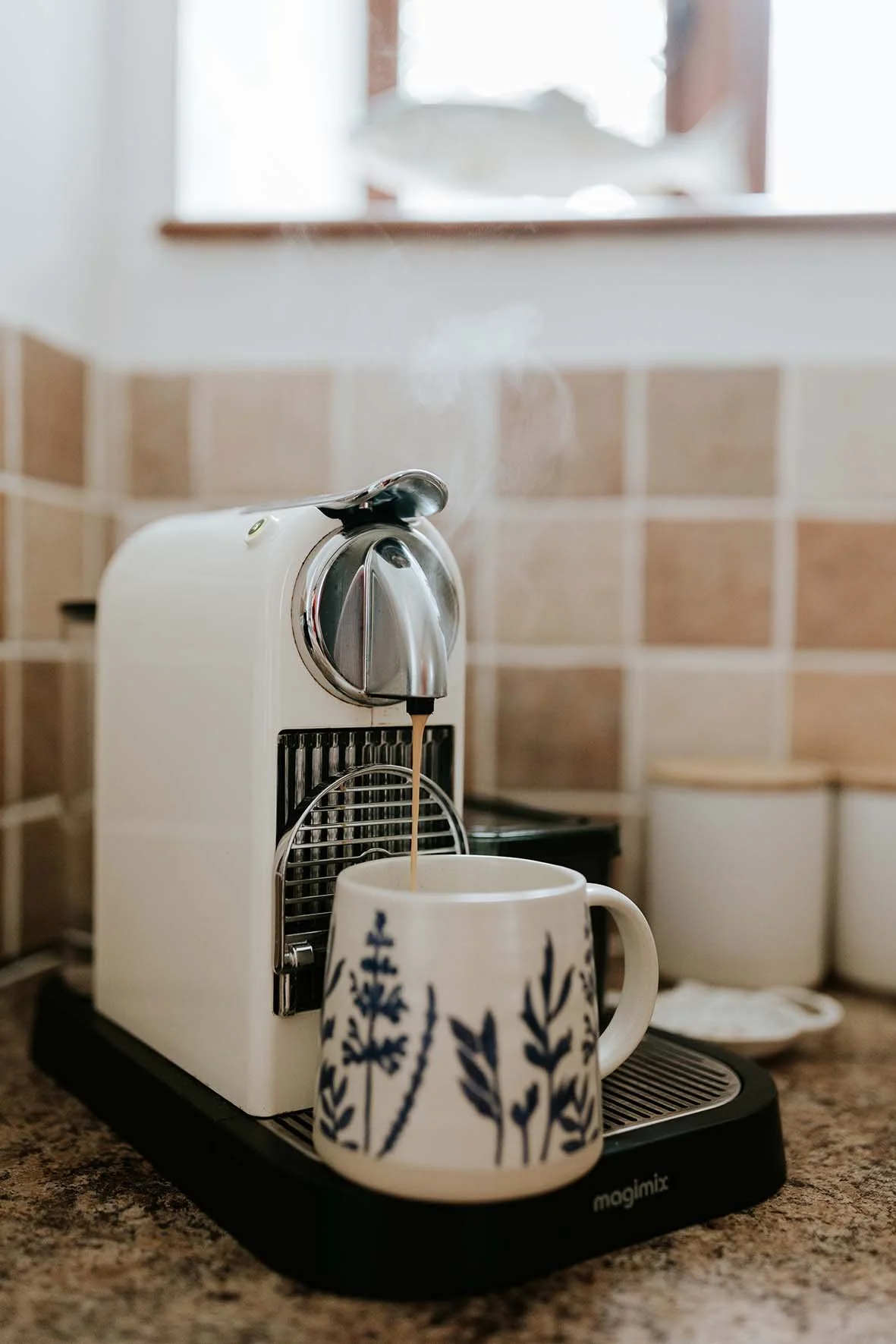 Coffee brewing from a coffee machine into a mug with a blue leaf pattern on a kitchen countertop.