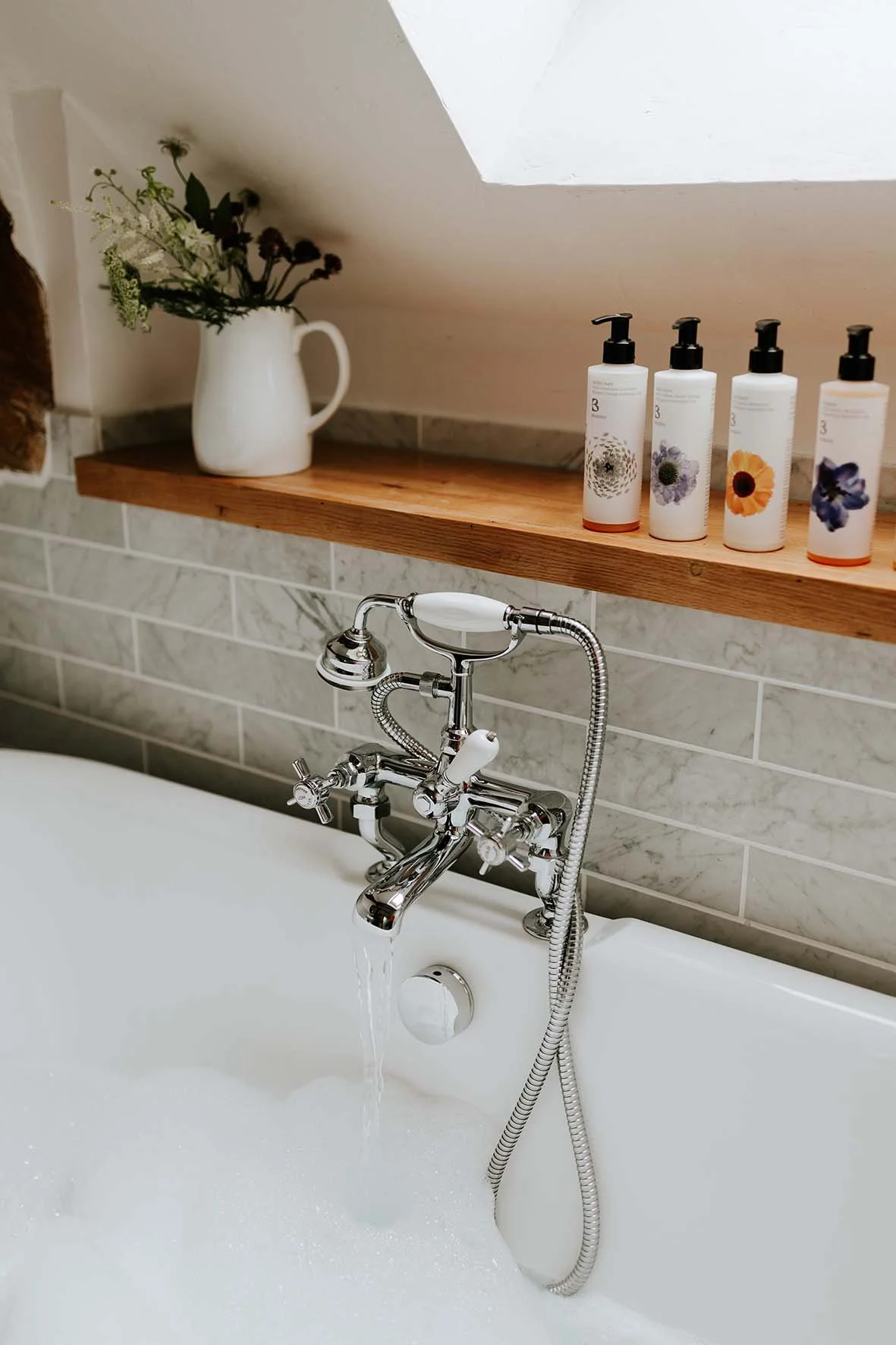 A white bathtub with a silver antique-style faucet and handheld shower head, filled with bubbly water. A wooden shelf above holds a white vase with flowers and four bottles of skincare or personal care products with colorful floral labels.