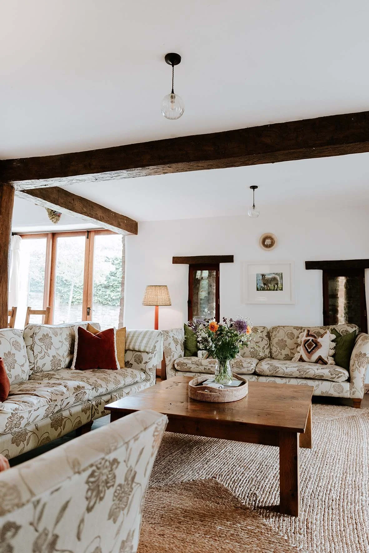 Living room with floral-patterned sofas, a wooden coffee table with a vase of flowers, a lamp, and large windows showing greenery outside, decorated in a cozy, rustic style.