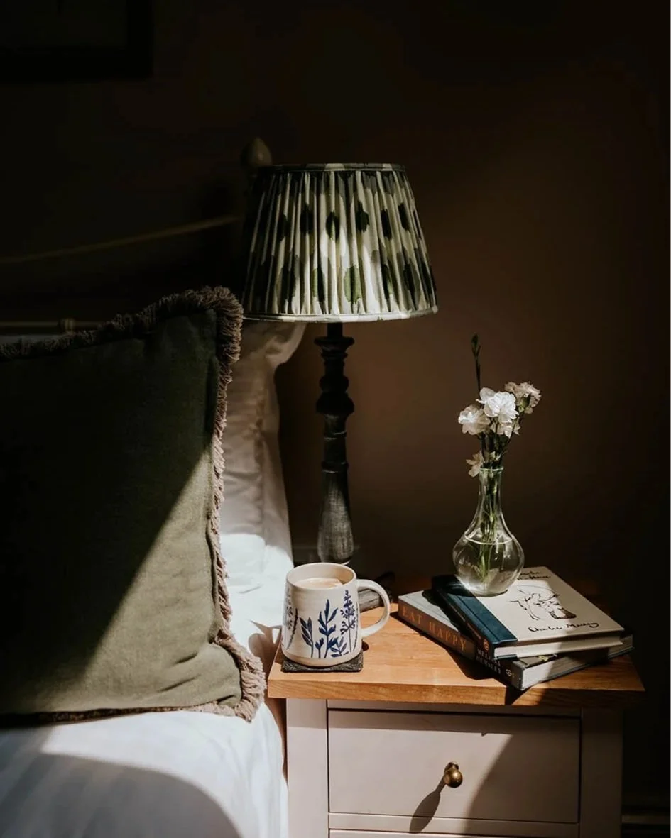 A bedside table with a cup of coffee, a stack of books, a glass vase with white flowers, a table lamp with a patterned shade, and part of a bed with a green pillow. The scene is dimly lit, creating a cozy atmosphere.