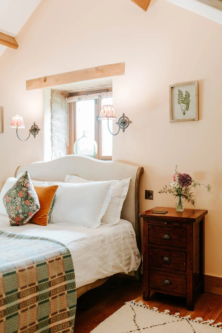 A cozy bedroom corner with a bed, pillows, a wooden nightstand with a vase of flowers, wall-mounted lamps, and a small window.