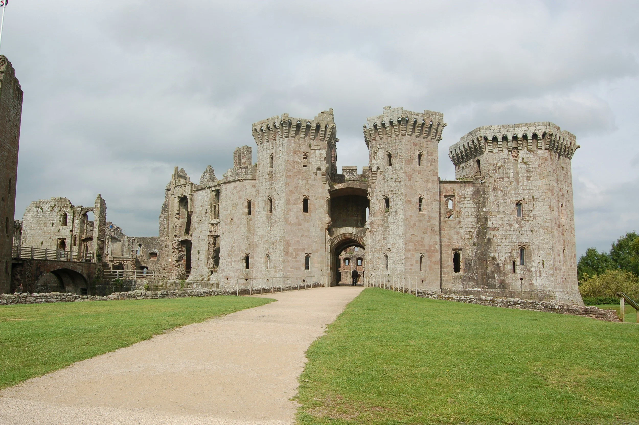 A medieval castle with multiple towers and a central gate, made of stone, surrounded by a grassy field and a pathway leading to the entrance.