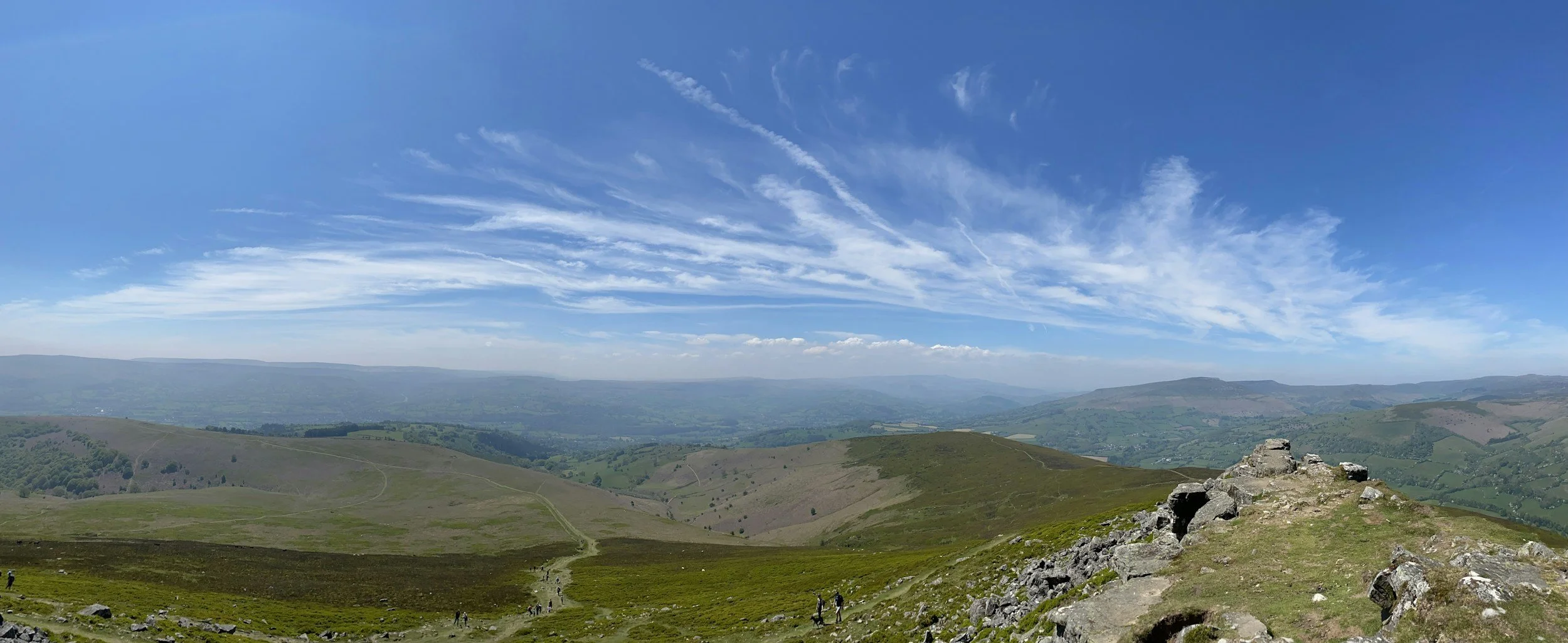 A panoramic view of rolling green hills under a bright blue sky with wispy white clouds and a few contrails, with hikers walking along a trail in the foreground.