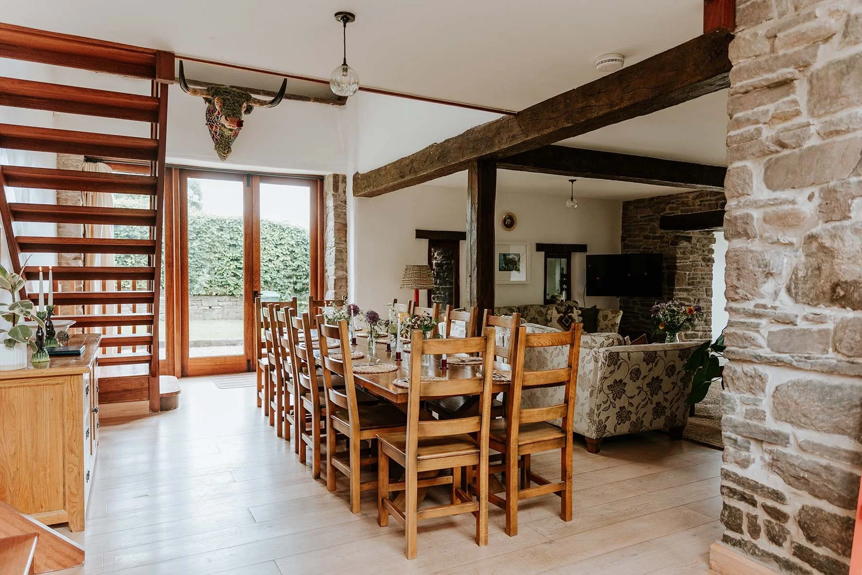 Dining room with a long wooden table, twelve chairs, flowers, and vases, opening to an outdoor patio. Interior features exposed wood beams and stone walls.