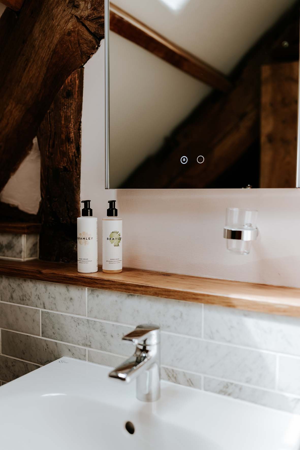 Bathroom sink with two white soap bottles labeled 'Bramley' on a wooden shelf, a mirror above, and wooden beams on the ceiling.
