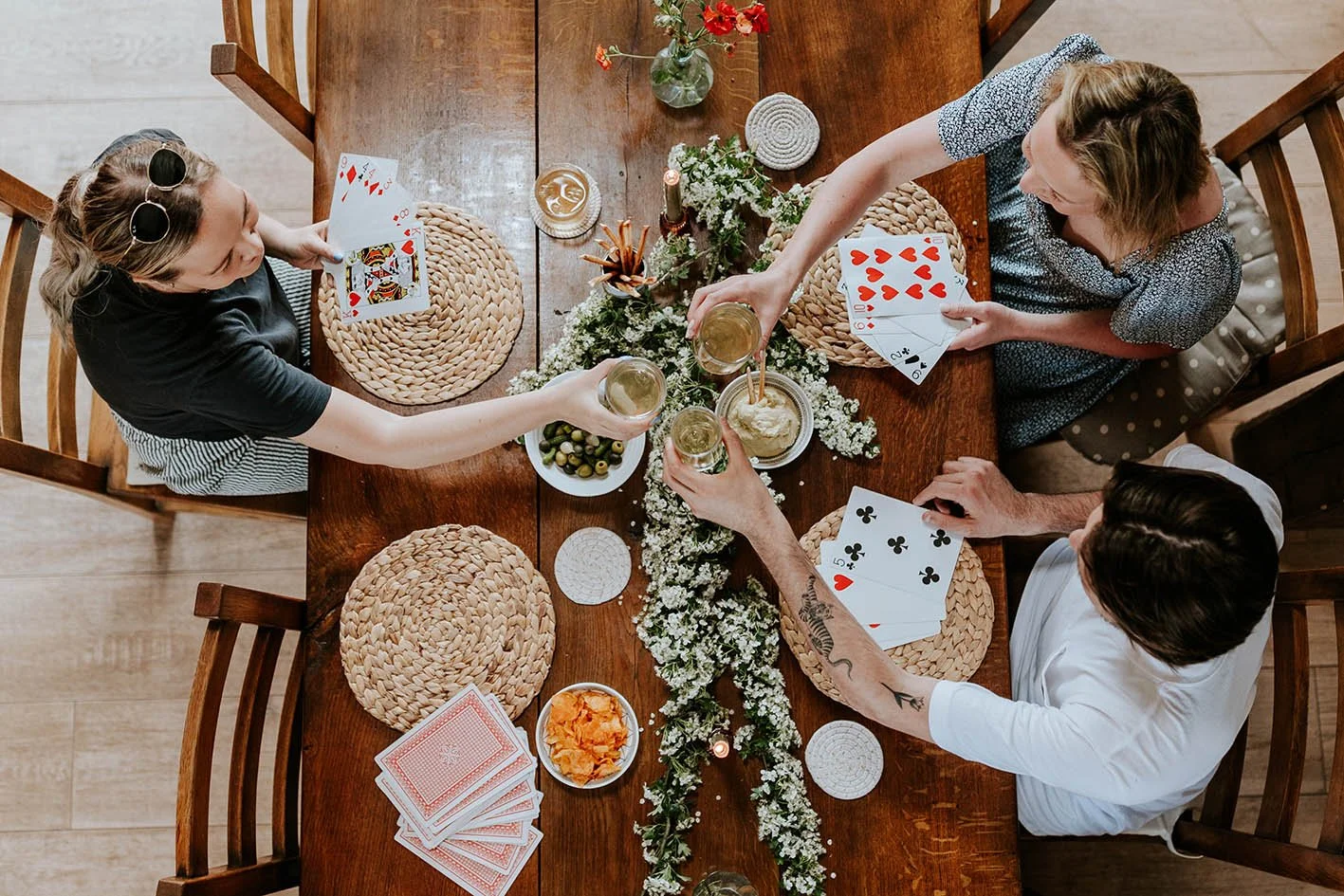 Three women playing cards around a wooden dinner table decorated with flowers and candles, with snacks and drinks present.