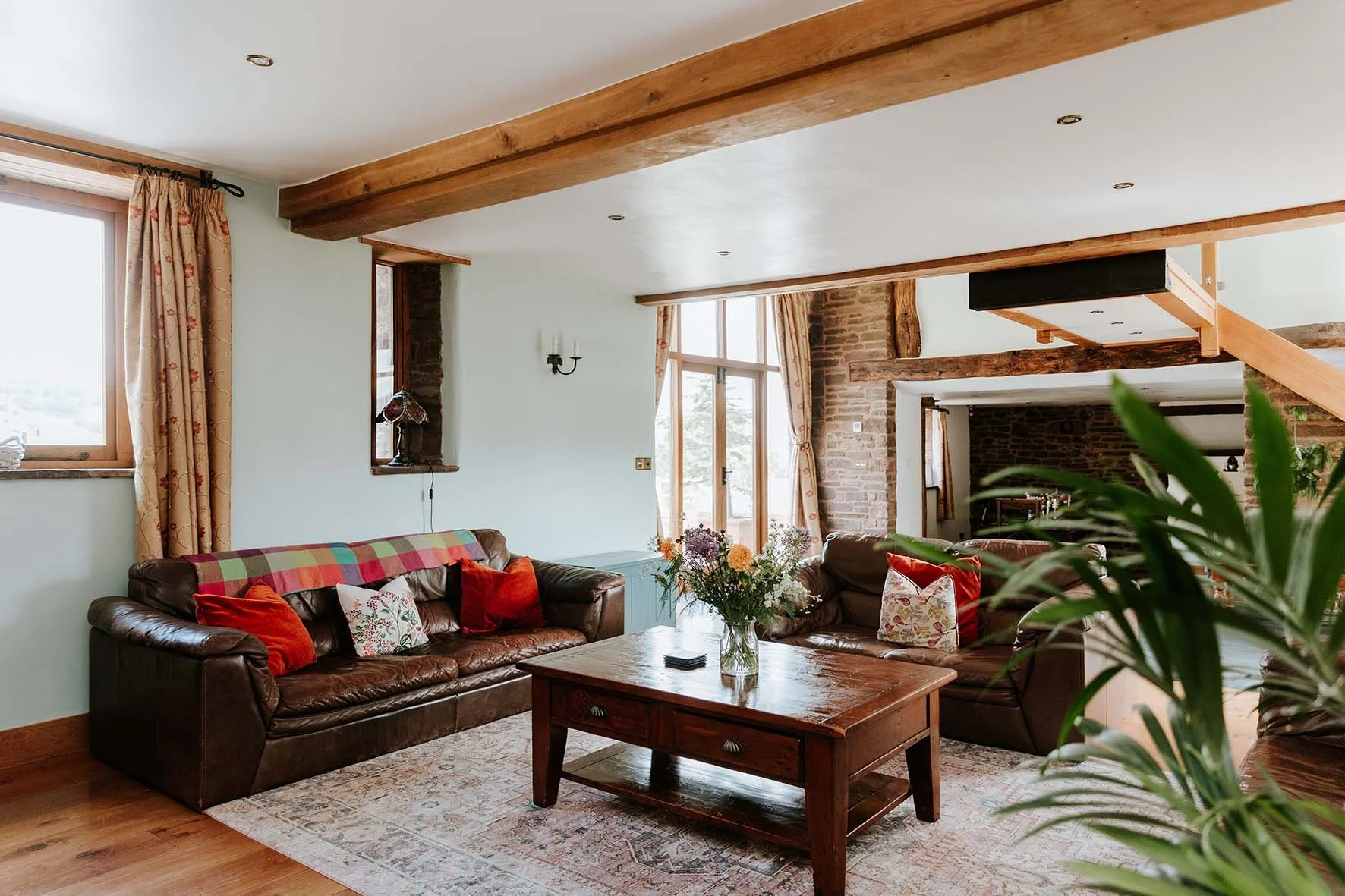 Cozy living room with wooden beams, brick accents, leather sofas with colorful cushions, a wooden coffee table, large windows with curtains, and plants.