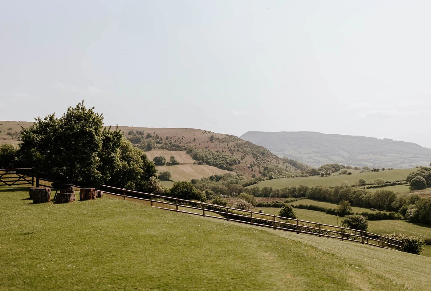 Open grassy field with trees, benches, and rolling hills in the distance under a cloudy sky.