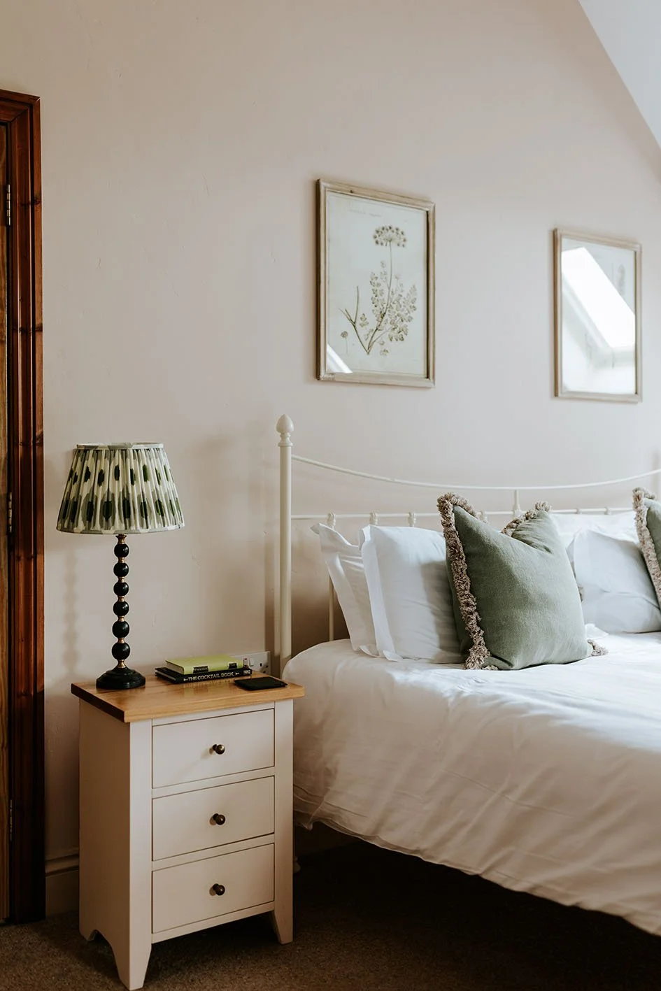 A cozy bedroom with a white bed, green pillows, a white nightstand, a black lamp with a patterned shade, and framed botanical prints on the wall.