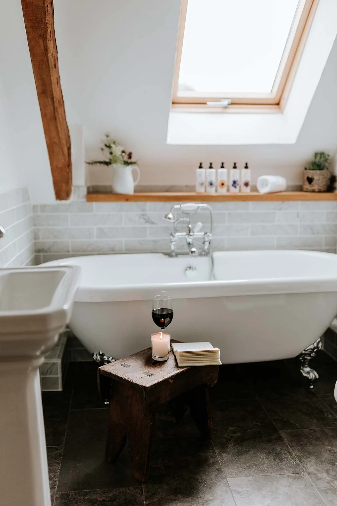 A cozy bathroom with a vintage clawfoot bathtub under a skylight, decorated with a small candle, a glass of red wine, and a stack of books on a rustic wooden stool.