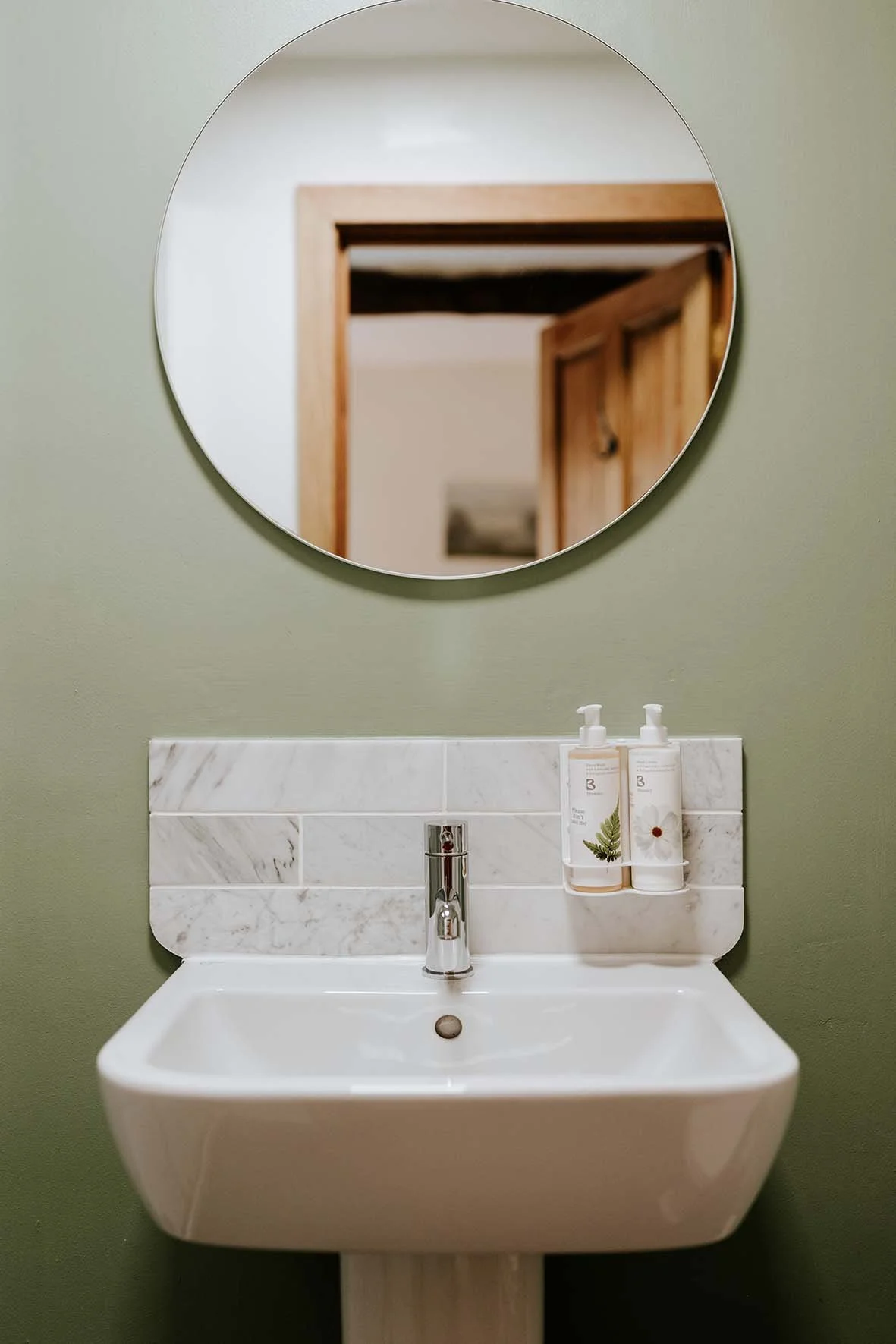 A white wall-mounted bathroom sink with a chrome faucet, above which is a rectangular marble backsplash, with two hand soap bottles placed on it. Above the sink is a round mirror reflecting a wooden door and part of a hallway.