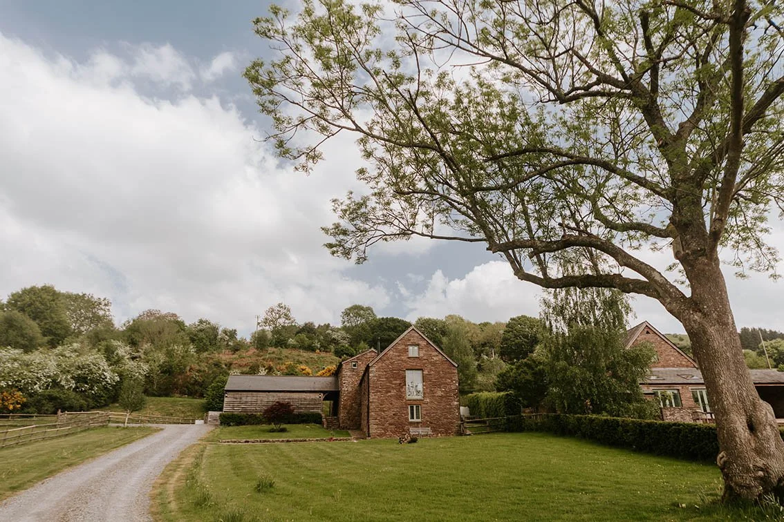 A rural scene with a gravel driveway, a brick house, a large tree, a well-maintained lawn, and surrounding greenery under a partly cloudy sky.